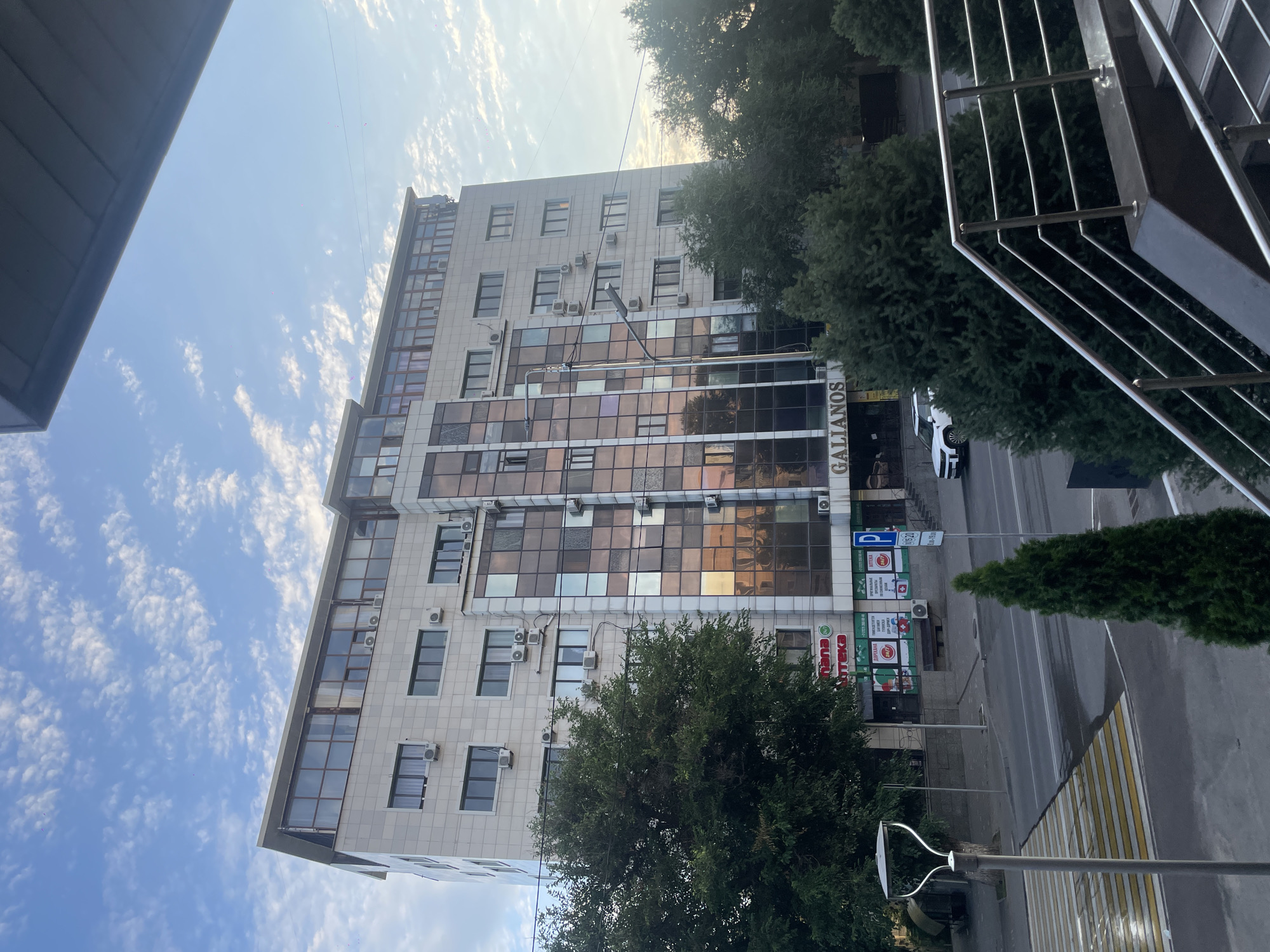 The image depicts the exterior of a modern, multi-story building situated on a street in Almaty, Kazakhstan. The building, identified by the signage "GALIANOS," features a light-colored facade with numerous windows, some of which are tinted and reflect the sky. The upper floors have a more modern design with extensive glass enclosures. Air conditioning units are visible mounted on the exterior walls.
In front of the building, a street with a crosswalk is visible. There is a lamppost near the crosswalk. Several trees line the street, some of which are tall and coniferous, while others are deciduous and fuller. A white SUV is parked or driving on the street.
Further details are visible at the ground level of the building, including signage for a pharmacy ("Аптека") with 24/7 service displayed. There are also signs indicating parking, and some pedestrian stairs with metal railings are present in the foreground, suggesting an entrance or elevated walkway. The sky above is blue with scattered, wispy clouds, indicating daytime. The overall impression is an urban street scene in a developed area.