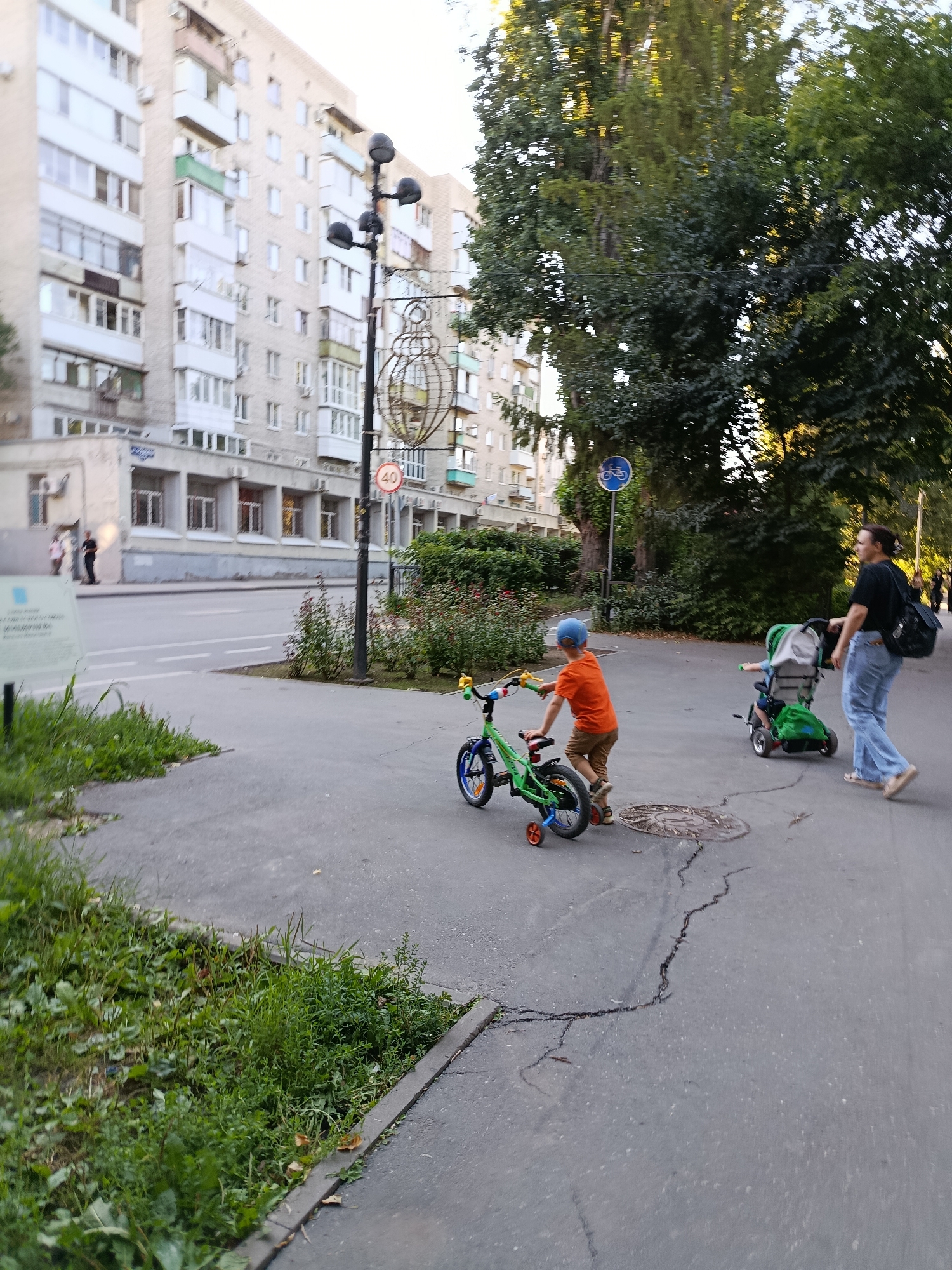 The image depicts a street scene in Saratov, Russia, on a clear day, likely late afternoon, judging by the warm sunlight filtering through the trees. In the foreground, a young boy wearing an orange t-shirt and a blue helmet is riding a small green bicycle with training wheels. He appears to be focused on his ride, moving along a paved sidewalk.
Behind him, a woman in a black t-shirt and blue jeans is walking, pushing a green stroller. The stroller appears to have a child in it, though the child is not clearly visible. The woman is looking ahead, suggesting she is accompanying the child on the bike or heading to a destination.
The setting is urban, with a multi-story apartment building visible in the background. The building has a light-colored facade with numerous windows and balconies. The street in front of the building is paved, and a sidewalk runs alongside it. There are also trees and bushes bordering the sidewalk, adding a touch of nature to the urban environment.
Several signs are visible. A circular sign with a red border and the number "40" is mounted on a lamppost. Another blue circular sign with a bicycle symbol is also present. A white informational sign is partially visible on the left side of the image.
The overall impression is a peaceful, everyday moment of a family enjoying an outdoor activity in a residential area. The sunlight and the presence of children suggest a pleasant, relaxed atmosphere. The pavement shows some cracks and wear, typical of urban infrastructure.