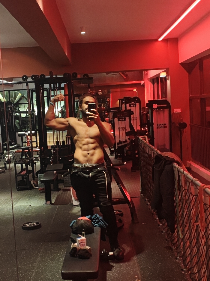 A male individual is captured in a full-body selfie within a gym setting, identified as located in the Chongqing High-tech Zone, China. The man, shirtless and appearing to have a lean, muscular physique with visible abdominal definition, stands facing a mirror, holding a smartphone in his right hand to capture the image. His left arm is flexed, displaying his bicep. He has long, dark hair tied back, a beard, and wears dark track pants with white stripes, along with an ear piercing and a wristband on his left arm.
The gym interior is characterized by strong red lighting from strip lights and possibly other sources, creating a distinct ambiance. Various workout equipment is visible in the background and foreground, including weight racks, benches, and resistance machines. Notably, "HAMMER STRENGTH" branding is discernible on at least two large machines to the right of the subject. The floor is covered with black rubberized gym matting. To the immediate left of the subject, a black weight bench holds a black baseball cap, a blue fabric item, and a small white carton beverage with a straw. In the mid-ground, a black safety net or barrier is positioned along a railing. Elements such as exposed ductwork or pipes are visible in the ceiling area. Reflections of the gym environment and the subject are visible in the mirrored surfaces.