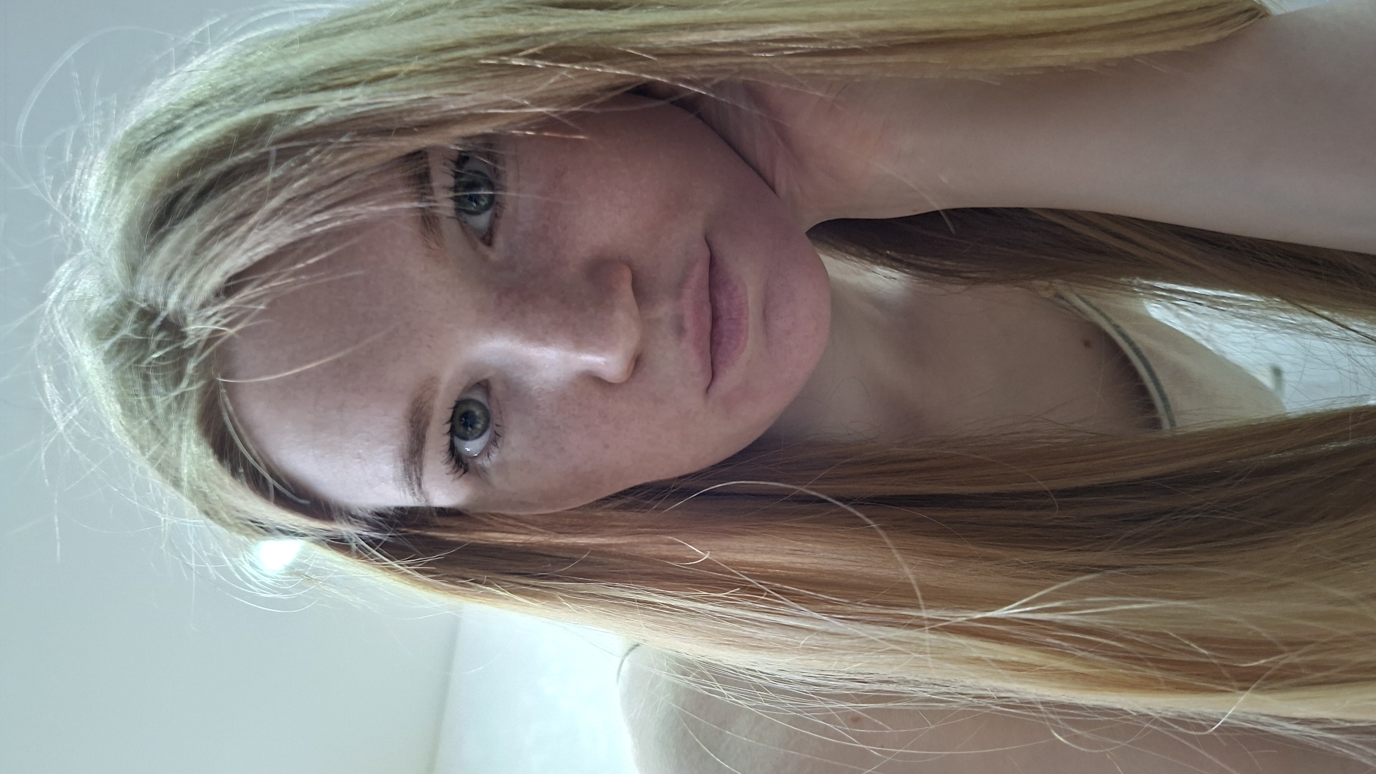 A close-up selfie captures a fair-skinned woman with light blonde and red-toned hair framing her face. Her features include freckles, light eyes, and full lips. She is wearing a light-colored top with a thin strap visible. The background appears to be a plain, light-colored surface, possibly a wall or ceiling. There is no discernible indication of the location being the city of Chaikovsky, Russia, or any other specific city.