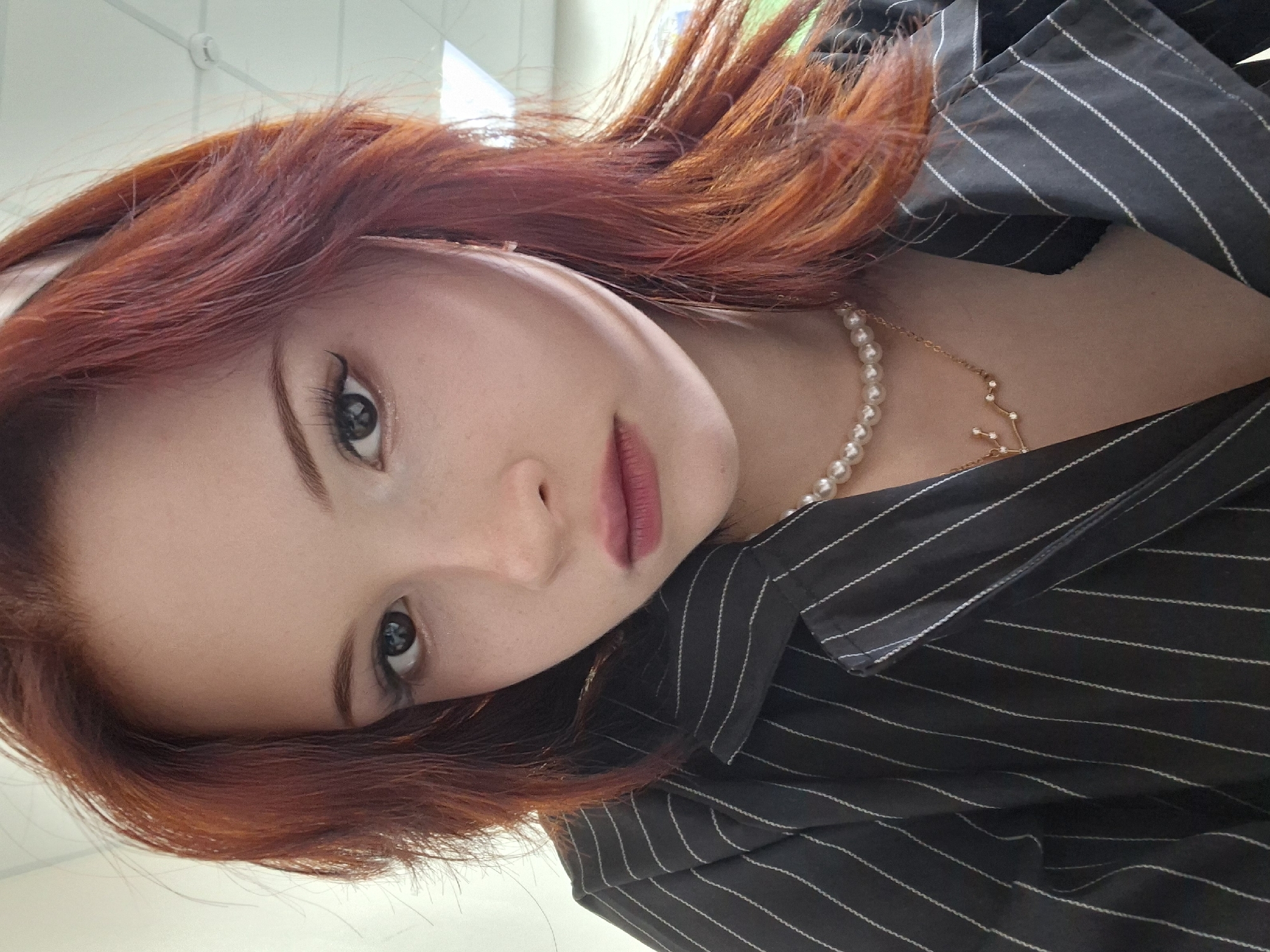 A portrait of a woman with red hair, wearing a black and white striped shirt, and a pearl necklace. The woman is indoors. There is no information to confirm that the location of the photo is Chaikovsky, Russia.