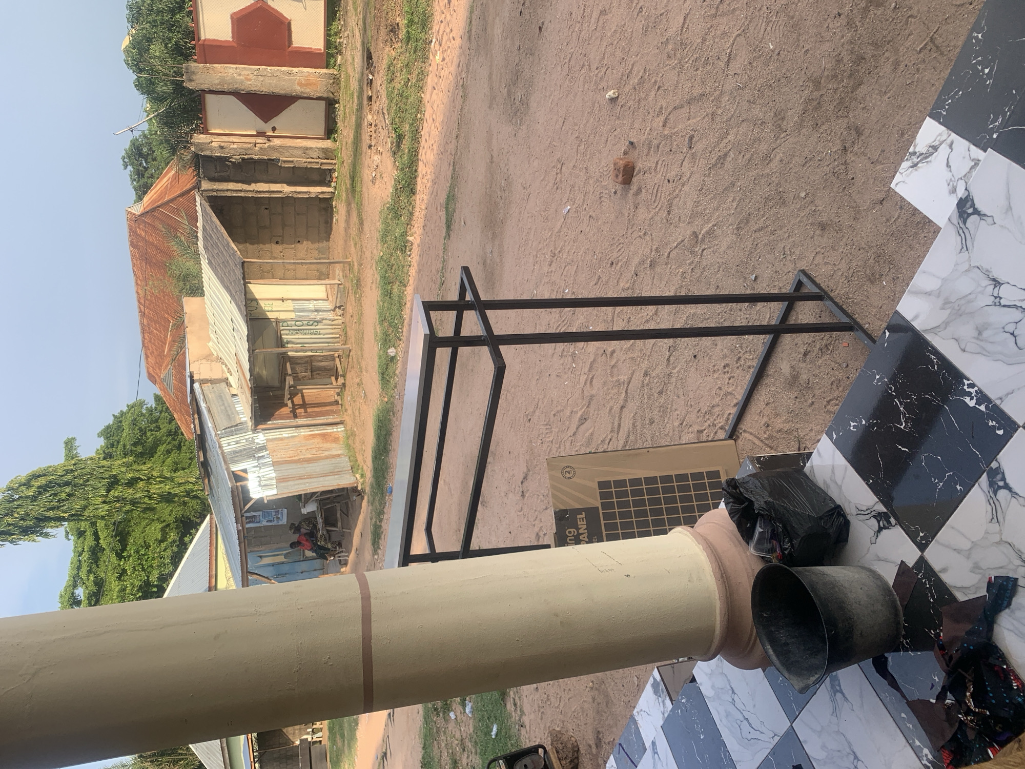 The image shows a street scene in Jauro Yinu, Nigeria. In the foreground, a large cream-colored column with a brown stripe is on the left, partially obscuring the view. In front of the column, there is a section of tiled flooring with a black and white marble pattern. To the right of the column, a black metal clothing rack with a light-colored top stands on the sandy ground. Leaning against the tiled flooring near the column are a dark grey bucket and a black plastic bag. Behind the clothing rack, a large rectangular object, possibly a solar panel, with the text "ing PANEL" visible, is propped up. The ground is sandy and appears to be a dirt road, with scattered debris and footprints. In the middle ground, there are several small structures with corrugated metal roofs and walls made of brick or wood. Two individuals are seated at a table under one of these structures. Further in the background, there are more buildings with reddish-orange tiled roofs and a tall, slender tree. A wall with a large red and white hexagonal pattern and a gate is visible on the right side of the background. The sky is clear and light blue, indicating it is daytime, likely morning or late afternoon due to the warm lighting.