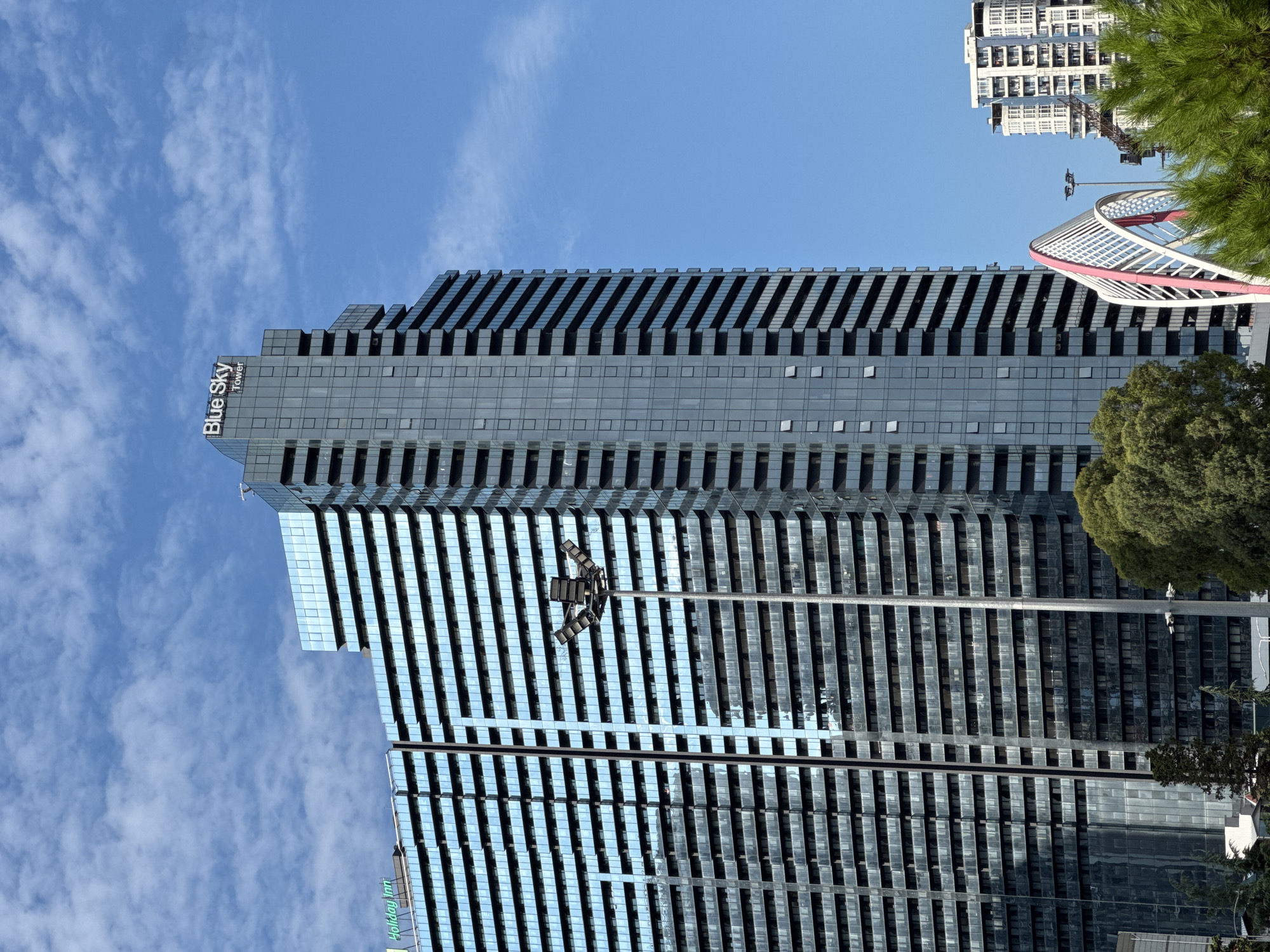 The image shows a view of the Batumi coastline in Georgia. The focal point is the Blue Sky Tower, a modern skyscraper with a dark glass facade. To the left, the Holiday Inn Batumi hotel is visible, also a tall building with a similar reflective glass exterior. In the foreground, lush green trees provide a natural contrast to the urban architecture. A white and pink arched structure, possibly part of a pedestrian bridge or decorative element, is also present. In the background, another residential building with white balconies can be seen. The sky is clear blue with scattered white clouds, indicating a sunny day.