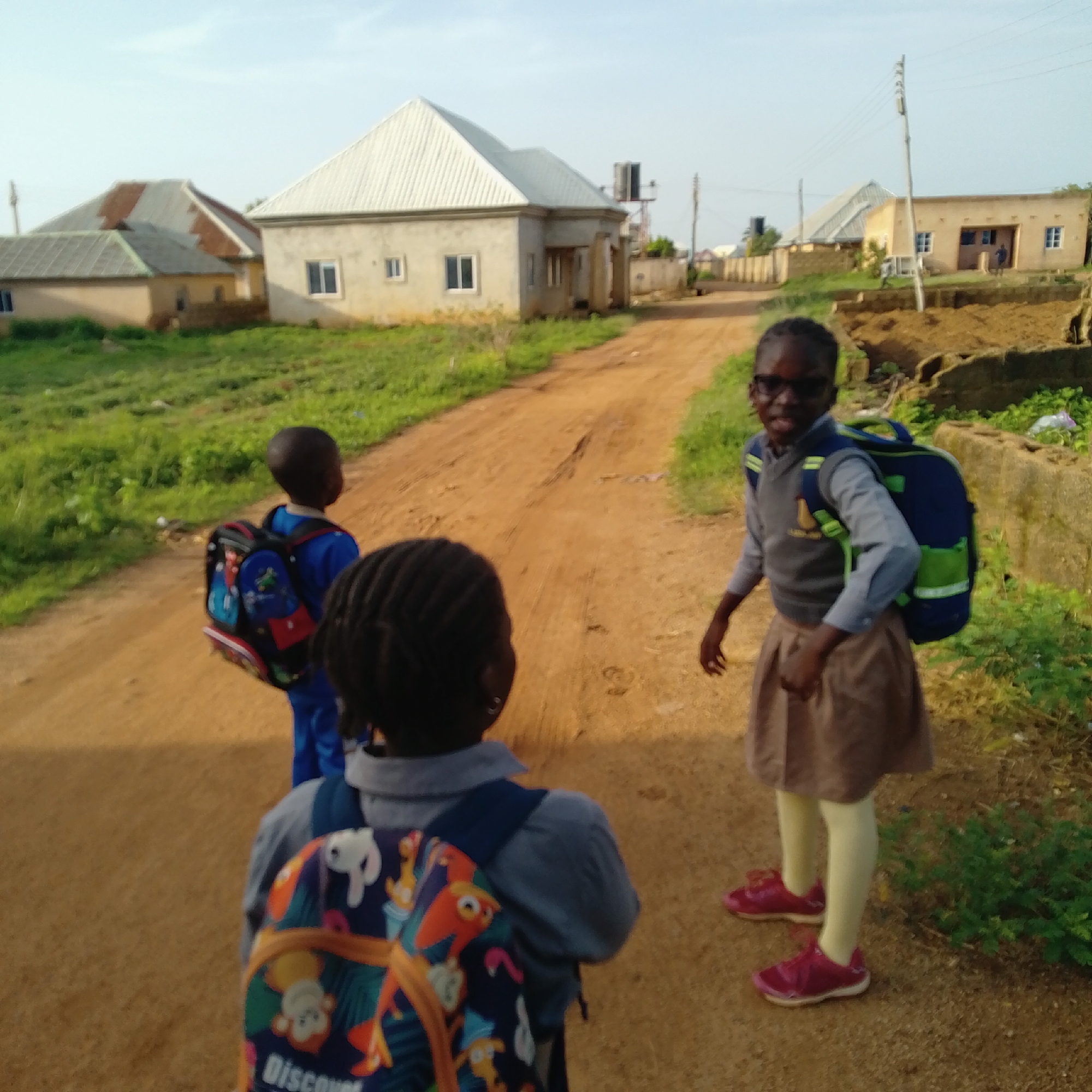 In Kwang, Nigeria, three children, two girls and one boy, walk along a dirt road. The boy, wearing a blue uniform, carries a backpack adorned with cartoon characters. One girl, also in a uniform and wearing glasses, sports a blue and green backpack. The other girl, facing away, has a colorful backpack. The scene is framed by a grassy verge and residential buildings with metal roofs. A utility pole stands to the right.