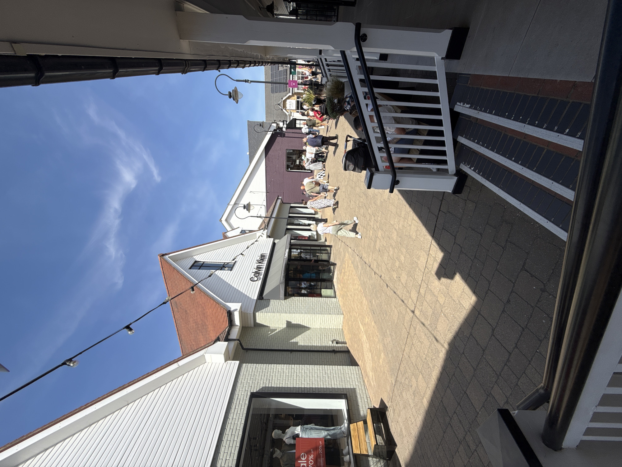 The image depicts an outdoor scene at a shopping outlet in Braintree, United Kingdom, likely during daylight hours with a clear blue sky and sparse clouds. The architecture suggests a retail environment, with multiple storefronts visible. One prominent store is identified as "Calvin Klein."
In the foreground, a dark railing and steps lead up to a white railing and a seating area where individuals are seated. The ground is paved with interlocking bricks. To the left, a store window displays mannequins and sale signage.
Further into the scene, several individuals are walking and browsing. Some are carrying shopping bags. A stroller is also visible. The environment appears to be a pedestrian walkway between shops. The overall impression is one of a busy, sunny day at a commercial district.