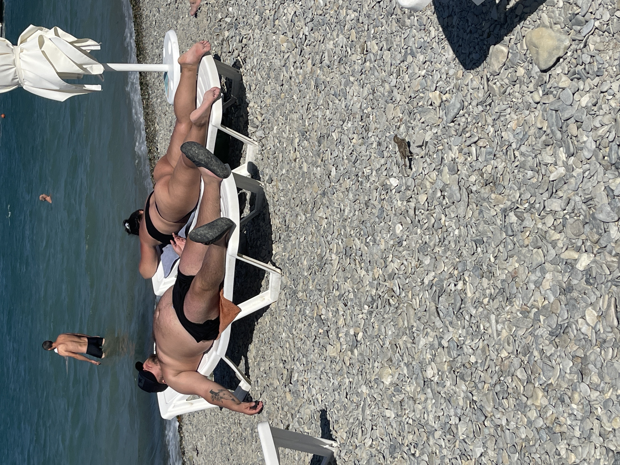 The image depicts a sun-drenched pebble beach in Gelendzhik, Russia, with the Black Sea as a backdrop. Two individuals are sunbathing on white lounge chairs. The person in the foreground, on the left, is a male wearing black swim trunks and a black baseball cap, with his arm outstretched. To his right, a female is lying on a lounge chair, also in black swimwear. In the water, a shirtless male stands waist-deep, and further out, a person can be seen swimming. A small bird is present on the pebble ground in the foreground. A white beach umbrella stands to the right of the lounge chairs. The scene is characteristic of a typical summer day at a Black Sea resort.