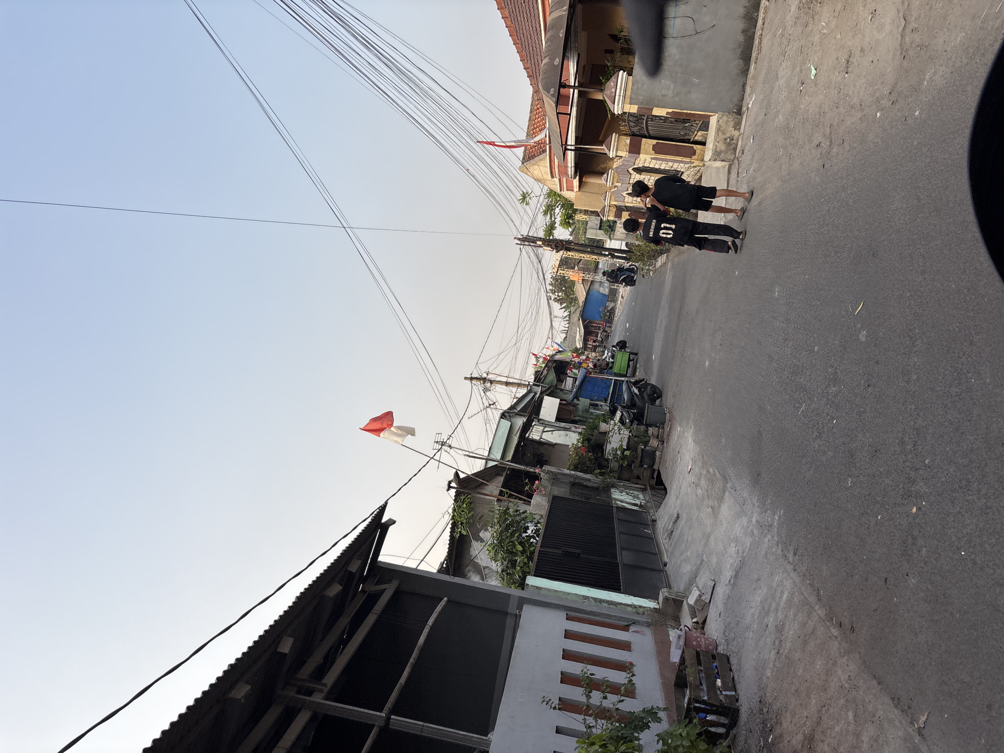 Two boys, one wearing a black t-shirt with "ANDHIKA 01" on the back, walk away from the camera down a narrow, paved street lined with houses and utility poles. An Indonesian flag flies from a pole on the right side of the street. The sky is clear and pale blue, suggesting it is late afternoon or early morning. The scene appears to be in a residential area within the city of Bekasi, Indonesia, characterized by its dense housing and overhead electrical wires.