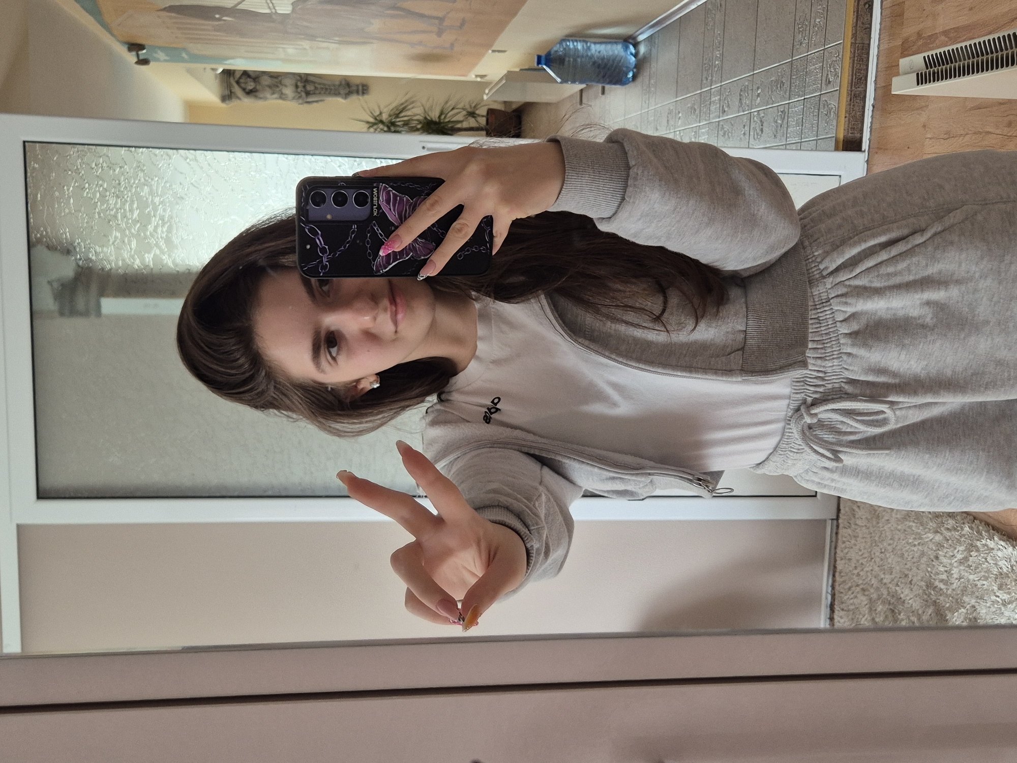 A young woman is taking a selfie in front of a mirror. She is wearing a grey sweat suit over a white t-shirt and making a peace sign with her right hand. She is holding a smartphone with a black case with a floral design. In the background, through a door, are a water bottle, plants, and tiled walls. The location is potentially in a house or apartment within Pavlikeni, Bulgaria.