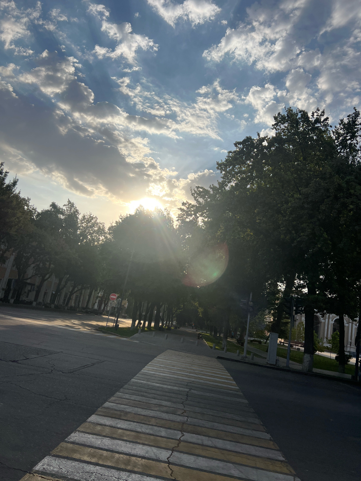 An eye-level, wide shot captures a sun-drenched street scene in a city, possibly during late afternoon or early morning. The sun, positioned high in the frame and slightly to the left, beams through dramatic clouds, casting rays of light across the sky and onto the landscape. A prominent crosswalk, with alternating white and yellow stripes, dominates the foreground, extending diagonally across the asphalt road. To the left of the crosswalk, the road is cracked and appears aged.
A row of lush, mature trees with dark green foliage lines the right side of the road, their branches forming a dense canopy that partially obscures the sky and buildings behind them. On the left side of the road, a sidewalk with a few scattered trees and a building with a light-colored facade are visible. The building appears to be a multi-story structure with windows and architectural details.
A red circular sign with a white horizontal bar, indicating "no entry" or a prohibition, is mounted on a pole to the left of the road, amidst the trees. Further down the road, a few blurry figures of pedestrians can be discerned, suggesting normal activity. The overall impression is one of a serene urban environment bathed in the warm glow of the setting or rising sun.