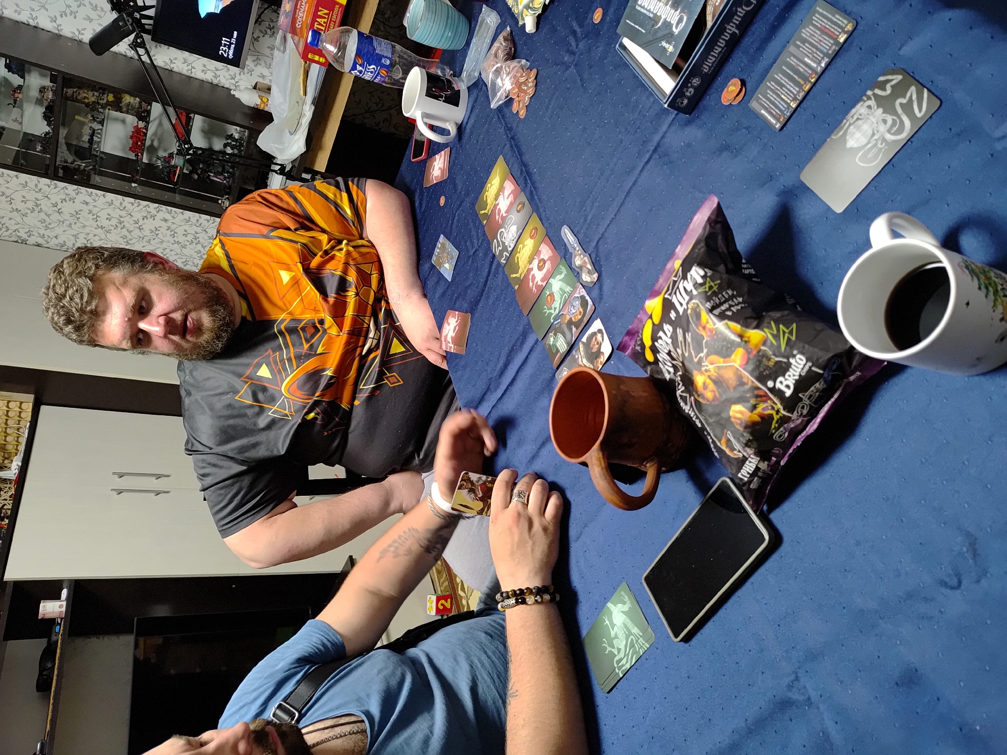 A medium shot captures two men playing a card game on a blue tablecloth-covered surface, possibly a table or large desk. The man on the left, with a full beard and wearing a colorful orange and black shirt, is looking at the camera. The other man, partially visible in the foreground, wears a blue shirt and has beaded bracelets on his wrist.
Scattered on the blue surface are various items: cards laid out in a row, chips in a plastic bag labeled with "Bruto" and the word "ГРИБЫ," two mugs with liquid, a ceramic mug, a mobile phone, a box with a game title that appears to be "Орифлаим," and scattered game tokens. The backdrop of the shot features a cabinet filled with figurines and display items, a television showing the time as 23:11 on Saturday, May 23rd, a desk lamp, and various packaged food items and bottles. The locale is likely a room in Марковский, Russia, inferred from Cyrillic text on the packaging.
