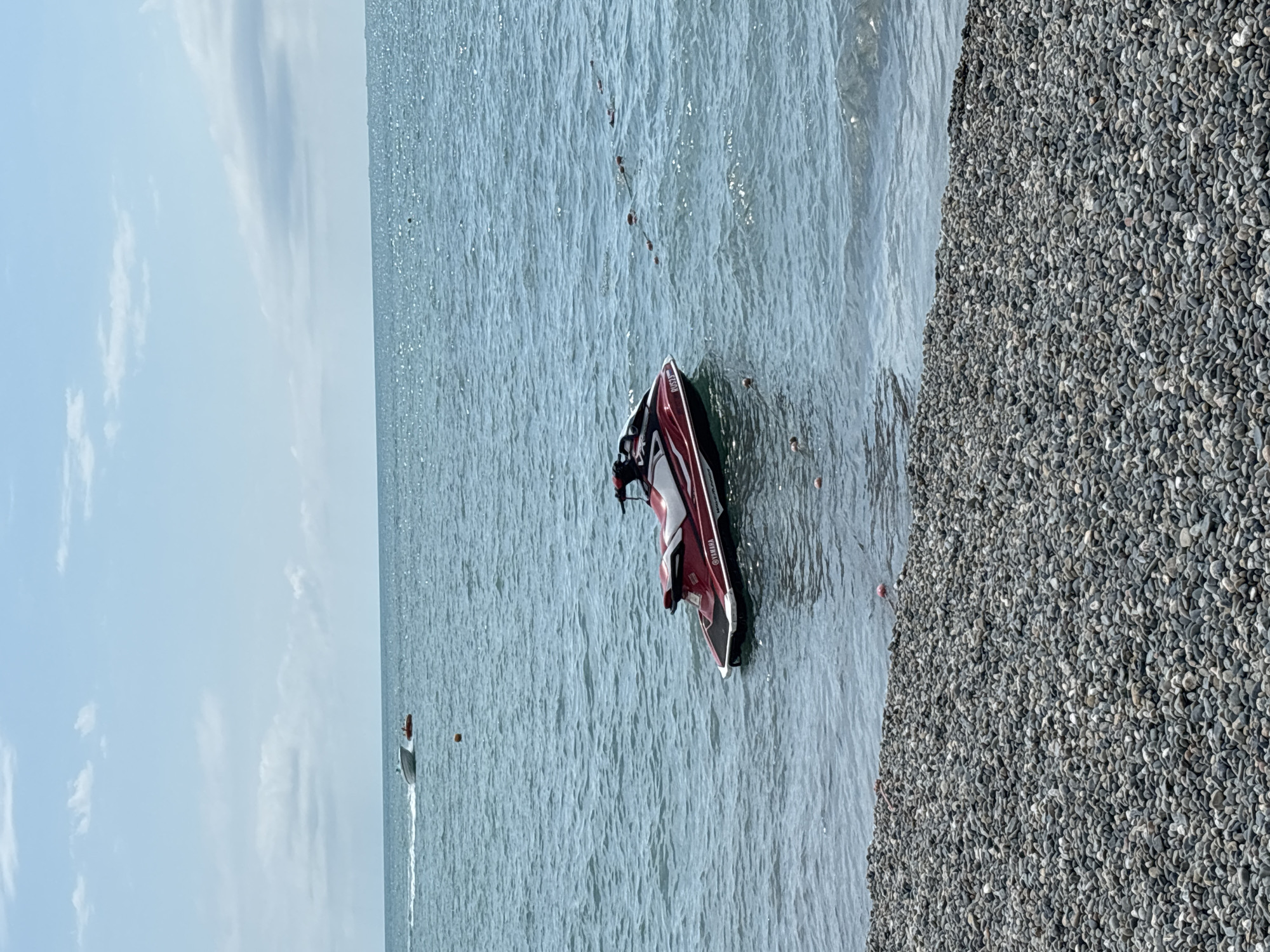 A red and white Yamaha WaveRunner personal watercraft is moored in the calm, blue waters of the Black Sea, close to a pebble beach. In the distance, a small white boat tows a red inflatable tube with a person on it, moving left to right across the frame. The sky is a pale blue with scattered white clouds. The scene appears to be on a beach in Sochi, Russia.