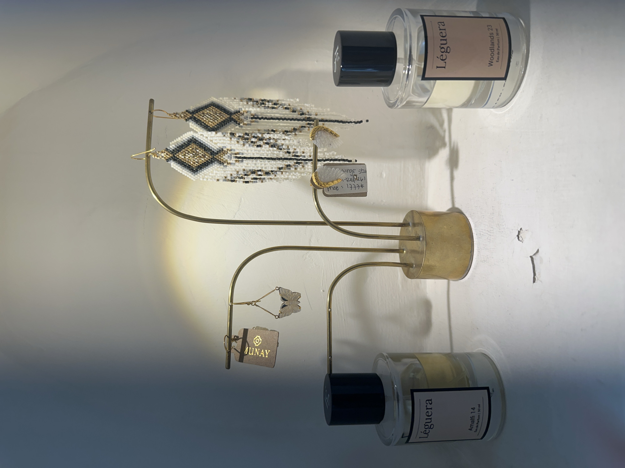 The image presents a display of artisanal goods. Dominating the scene is a gold-toned, multi-tiered earring stand. The top tier holds a pair of long, fringed earrings crafted with white, gold, and black beads, arranged in a geometric, possibly diamond-shaped, pattern. Adjacent to these are a pair of smaller, gilded earrings with white feather-like tassels. Dangling from a lower tier of the stand is a small tag with the word "MUNAY" printed on it, next to a butterfly shaped pair of earrings. Also attached is a small handwritten price tag "winge stu price: 1777* XI-REP 261. ". A cylindrical gold base supports the stand. Flanking the stand on either side are clear glass bottles of "Léguera" perfume, each with a black cap. One is labeled "Woodlands 23 Eau de Parfum | 50 ml", and the other "Amalfi 14 Eau de Parfum 50 ml". The composition is set against a stark white background. The location in North Kuta, Indonesia is not evident from the image.