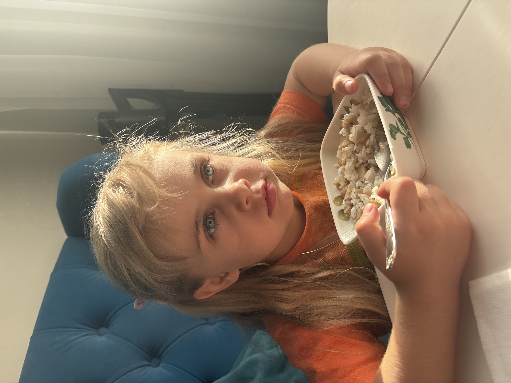 A young Caucasian girl with blonde hair and blue eyes sits at a white table, eating rice from a bowl with a spoon. She is wearing an orange t-shirt. She is seated in a blue upholstered chair. The lighting suggests it is daytime, with sunlight streaming in from the right side of the frame, casting shadows and highlighting the dust motes in the air. The background is blurred but shows curtains and possibly furniture. The setting is likely a residential interior. No specific location within Unknown, Russia, can be determined from the image.