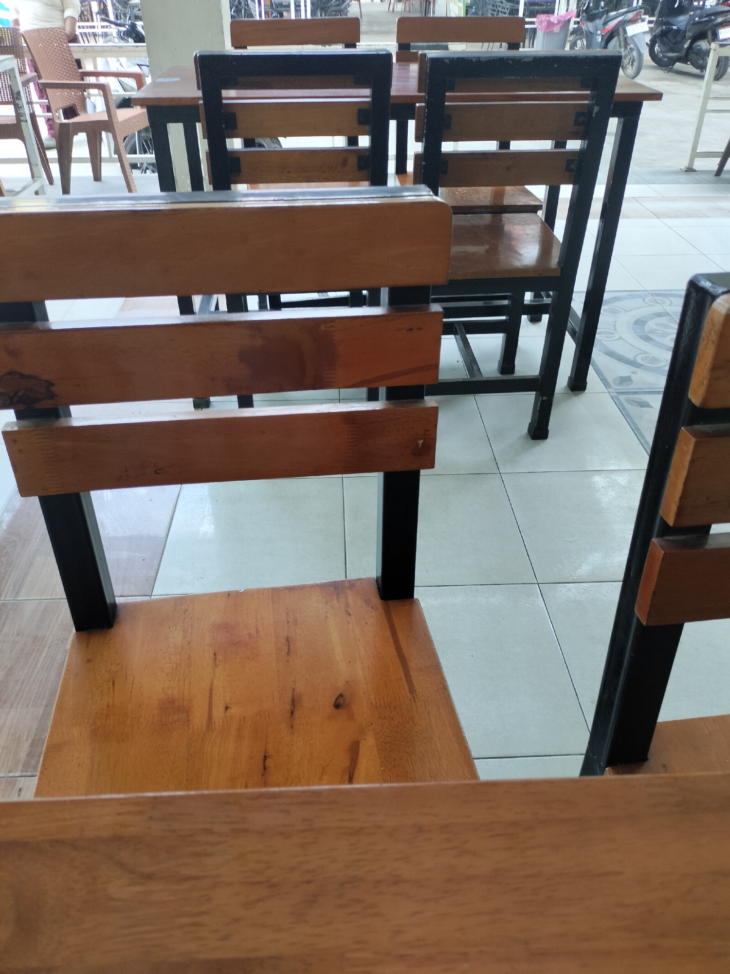 In Keudealue Ie Puteh, Indonesia, this image captures a close-up view of several wooden chairs with black metal frames. The chairs are arranged around tables, suggesting an outdoor dining or seating area. The foreground is dominated by the top of a wooden table or chair. In the midground, the backs and seats of three wooden chairs are visible, their slats and frames clearly defined. The background reveals more seating, tables, and a glimpse of what appears to be a street scene with motorcycles, indicating this is likely a public space such as a cafe or restaurant. The flooring is tiled in a neutral color, and the overall lighting suggests daytime.
