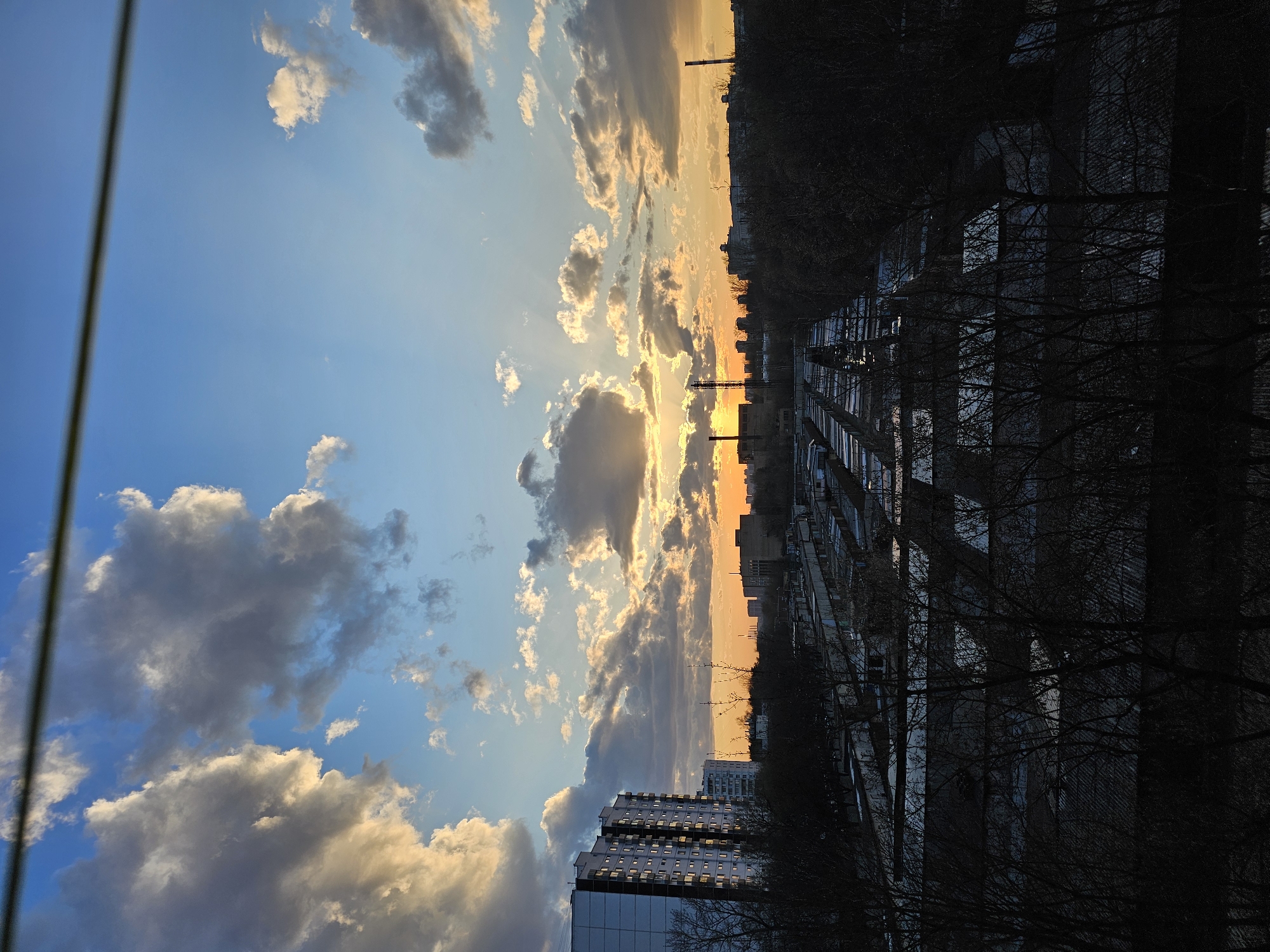 The image shows a sky with a blue expanse and puffy clouds illuminated by the setting sun, transitioning to a view of Khimki, Russia. The sun is positioned on the horizon, casting an orange glow between buildings and trees. A tall building is visible on the bottom left. Darker structures and trees occupy the foreground, creating a stark contrast against the bright sky.