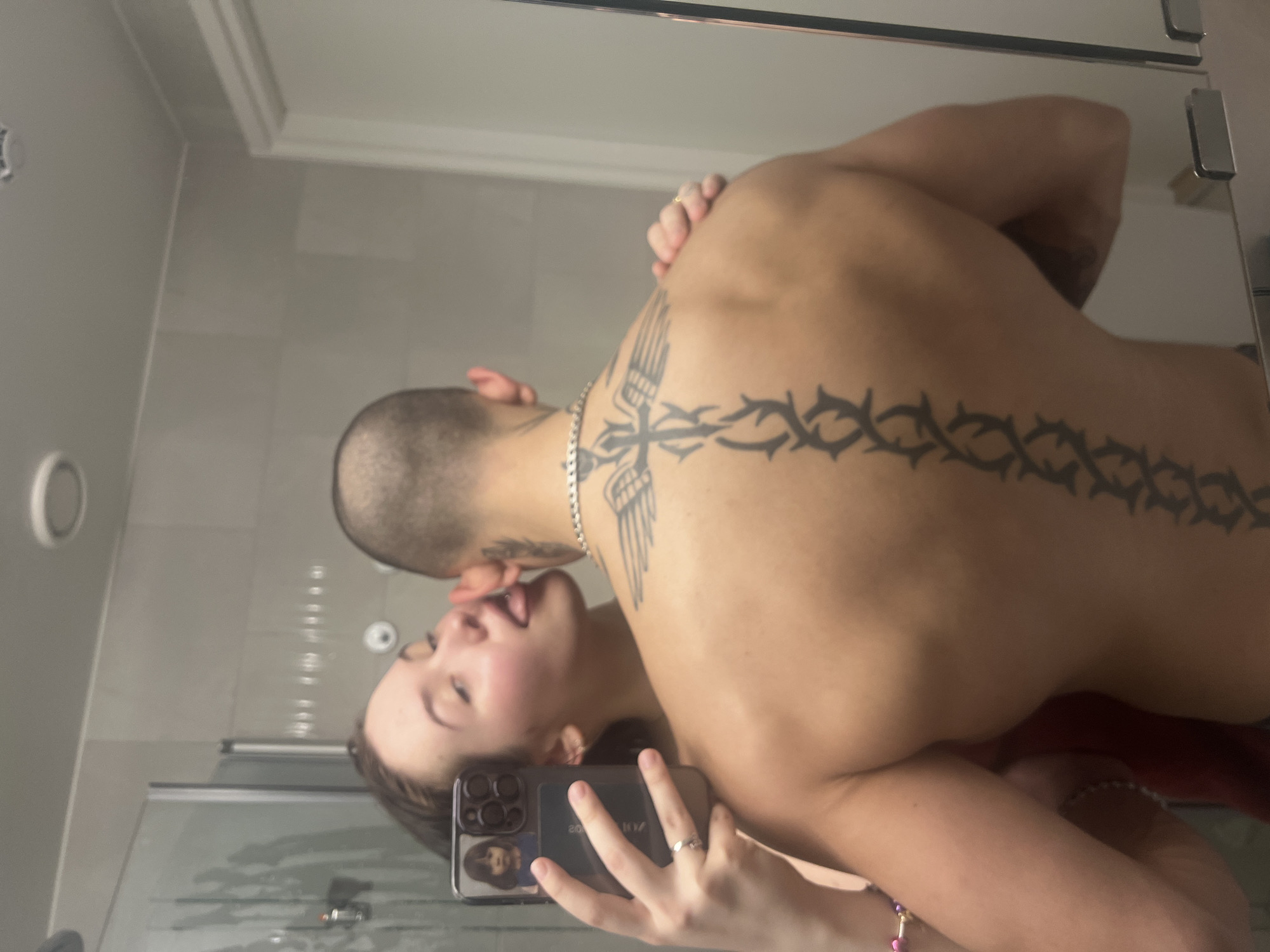 The image displays a mirror reflection of two individuals in what appears to be a bathroom in Vantaa, Finland. A female subject, partially visible on the left, holds a dark-colored smartphone in her left hand, taking a selfie. A small image of a child is visible on the phone's case. She wears a ring on her left ring finger and a multi-colored beaded bracelet on her left wrist. Her face is turned towards a male subject, and her tongue is extended, making contact with his ear/neck area. The male subject, positioned with his back to the mirror, has a shaved head and a muscular physique. He wears a silver-colored chain necklace. His back features a prominent tattoo depicting a winged cross design on the upper back, transitioning into a thorny, vine-like pattern extending down his spine. His right hand is visible resting on his upper back. The background shows light-colored tiled walls, a white ceiling with a circular light fixture, a glass shower enclosure on the left, and a white doorframe on the right.