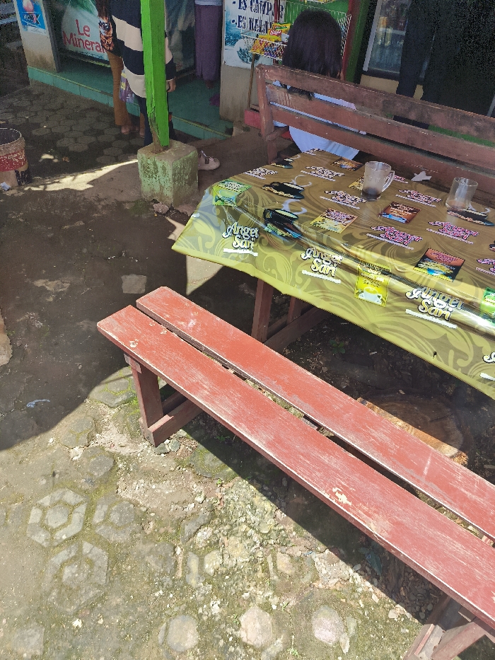 The image shows a red wooden bench with two seats, located in a seating area outside a shop in Majalengka, Indonesia. The coordinates provided, -6.846 latitude and 108.204 longitude, do not pinpoint the exact location but place it in the general vicinity of Majalengka, a municipality in the West Java province of Indonesia. The bench is positioned beneath a table covered in a yellow and green tablecloth with various colorful advertisements for a product called "Sari". The table is occupied by a person seated on a brown wooden bench, facing away from the camera. The seating area is located on a paved surface, which has grass growing between the stones. The environment is shaded, with the sun casting long shadows. The bench is old and weathered, with signs of wear and tear. There is a blue object on the ground near the bench, possibly a bag or other item. The image was taken from a low angle, highlighting the details of the bench and the tablecloth.