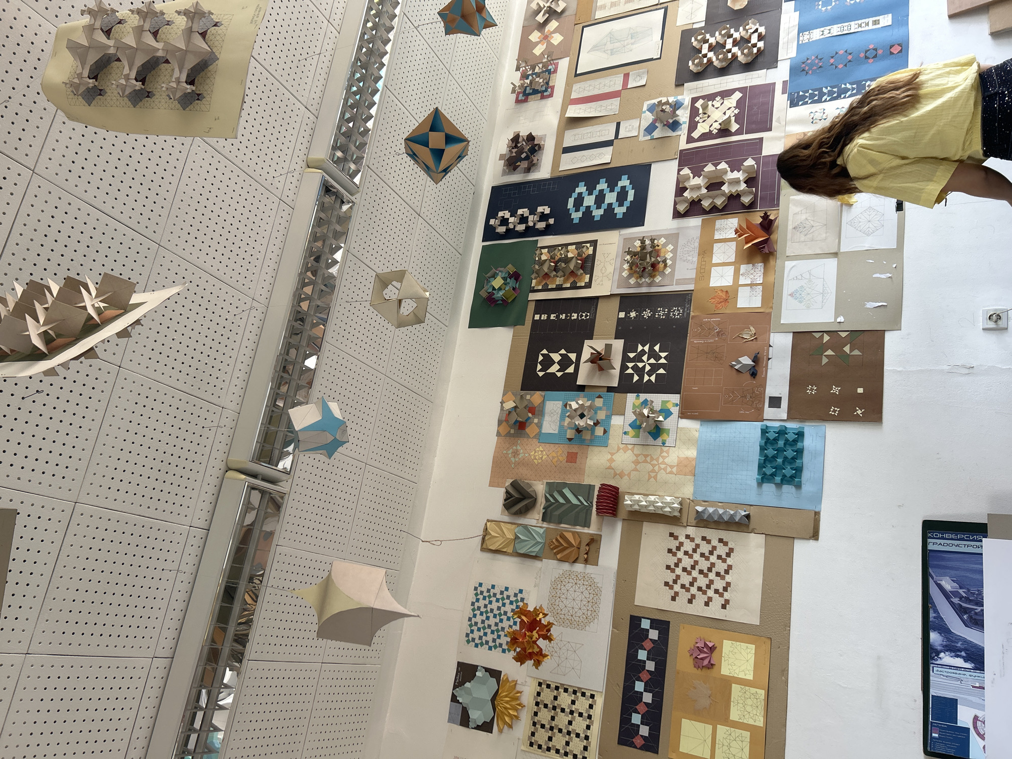 This image presents an upward-angled view of an indoor exhibition or display space, likely a design studio or gallery, located in Sofia, Bulgaria. The primary focus is a large white wall densely covered with various geometric artworks and architectural models. The ceiling, composed of white perforated acoustic panels, features several long, rectangular light fixtures with reflective, grid-like interiors.
The wall display is an eclectic collection of two-dimensional patterns and three-dimensional paper constructions, meticulously arranged on different colored backgrounds. The 2D elements include hand-drawn schematics, printed geometric patterns, and tessellations. These are interspersed with numerous 3D models crafted from paper or cardstock, demonstrating complex folds, modular designs, and relief structures. The models vary significantly, from intricate star-like shapes and interlocking cubes to more organic, wave-like, or fan-shaped forms, often displaying repetitive geometric motifs. Many are monochromatic, while others incorporate multiple colors.
Several lighter-toned, multi-faceted geometric shapes, possibly polyhedra or abstract forms, are suspended at different heights from the ceiling by thin wires. One distinct hanging piece features a two-tone blue and beige design.
In the upper right quadrant, a person's upper torso and head are visible, leaning into the frame from above. The individual has long, wavy brown hair, wears a yellow shirt, and dark patterned pants, appearing to be observing the display or possibly capturing an image.
On the far right, a partially visible framed poster with Cyrillic text provides a location cue. Key visible words include "КОНВЕРСИЯ" (Conversion) and "ГРАДОУСТРОЙ" (Urban Planning/Development), suggesting an academic or professional context related to architecture or urban design. A white electrical outlet is also visible on the wall in this area. The scene conveys an atmosphere of creative exploration and academic presentation through geometric art and design.