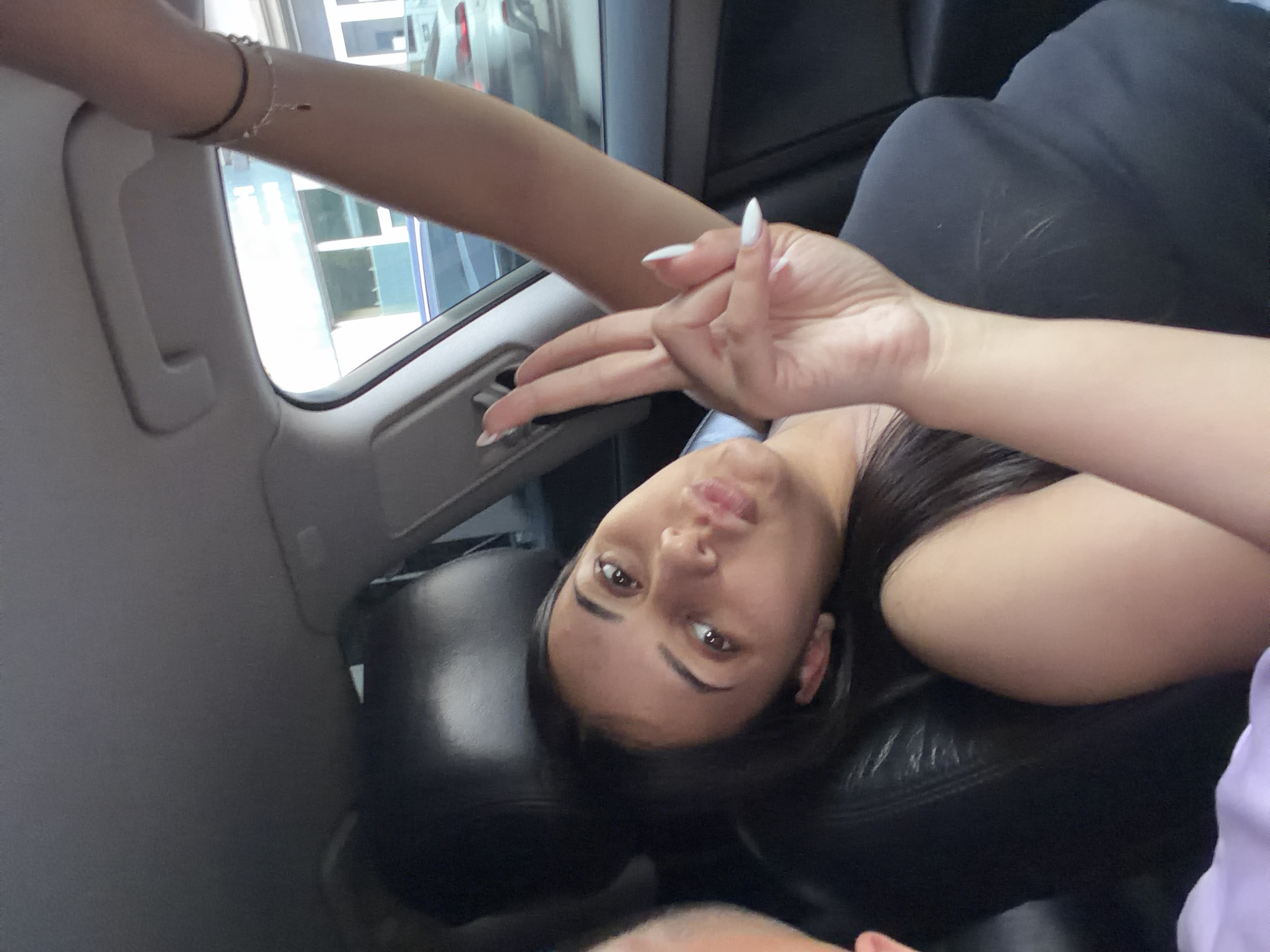 The image displays a young woman lying down or reclining in the interior of a car, looking directly at the camera. Her head is positioned at the lower center, slightly tilted to her right, showcasing her dark hair, dark eyes, and full lips. She is wearing a dark-colored top. Her right arm is extended towards the upper left side of the frame, adorned with a slender bracelet. Her left hand is visible on the right, performing a hand gesture with splayed fingers, highlighted by long, white manicured nails. The background reveals the car's interior, including a light grey ceiling panel and a window on the left. Through the window, blurred outdoor elements, possibly buildings or other vehicles, suggest it is daytime. A partial view of another person's head or body is visible at the very bottom of the frame. The scene takes place within a car, in Shumen, Bulgaria. No visible text is present.