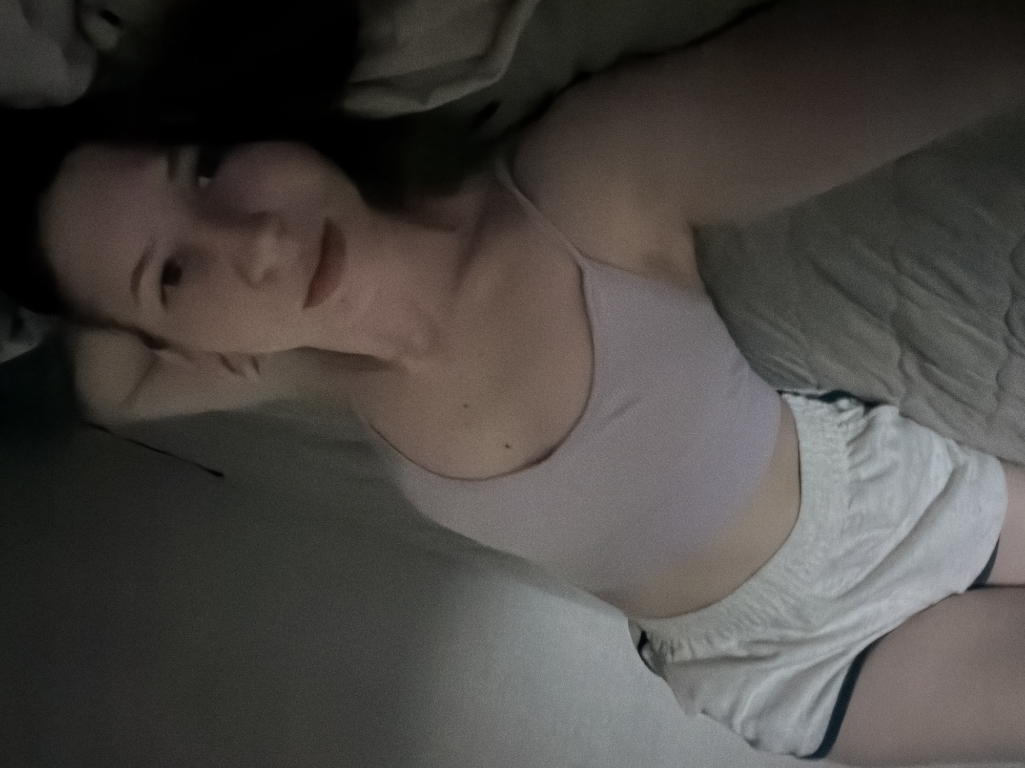 A person is lying on their side on a bed with a gray comforter. They are wearing light-colored shorts and a light-colored tank top. The person's face is partially visible, and they are looking towards the camera. The image appears to have been taken in a dimly lit room. There is no specific information available to determine the exact location within Dongducheon-si, South Korea.