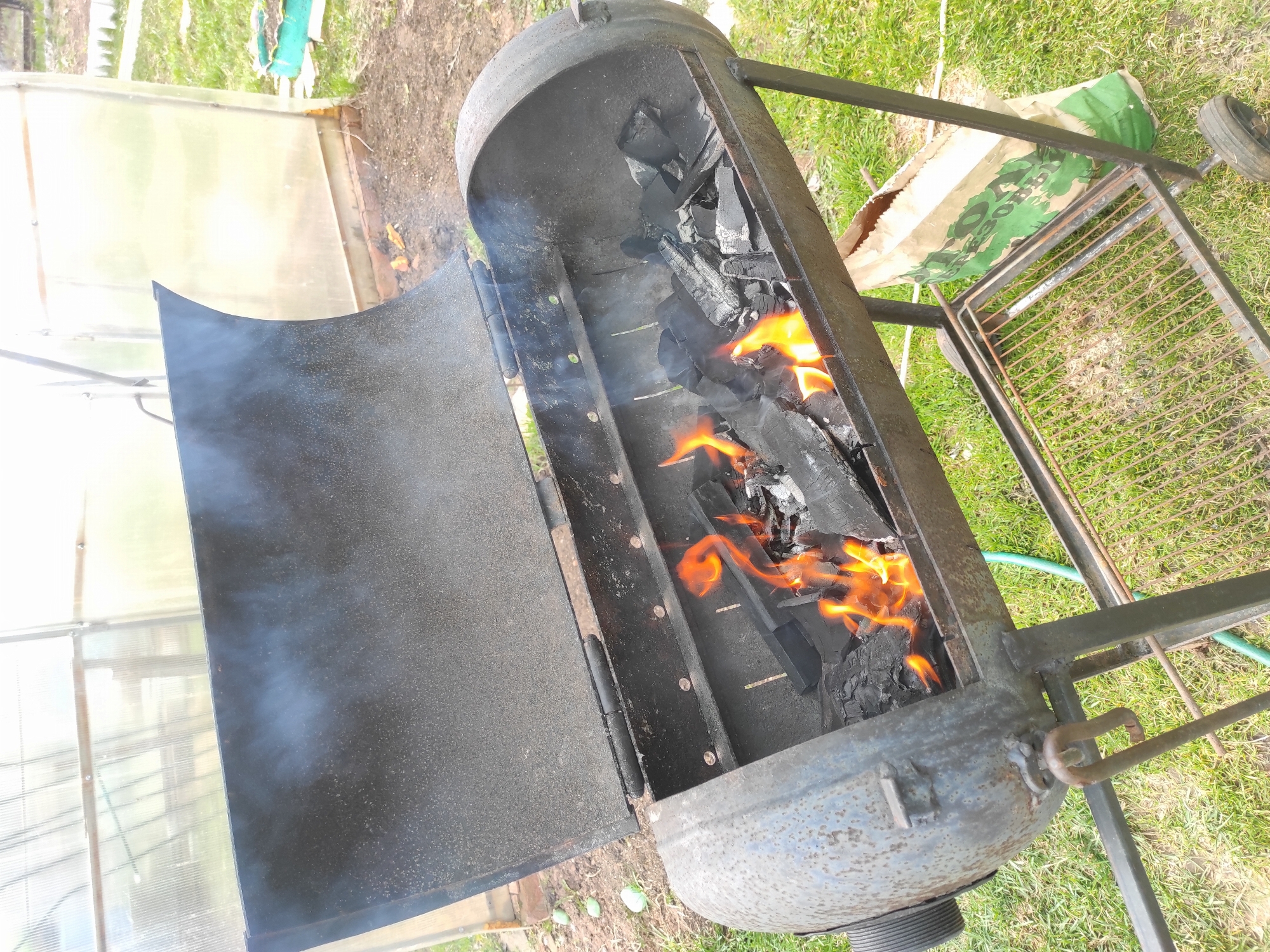 An outdoor scene features a robust, barrel-shaped barbecue grill, likely custom-made, with an open lid. Inside the grill, a mix of charcoal and wood is actively burning, producing vibrant orange flames and light grey smoke that drifts upwards. The grill is mounted on a metal frame, which includes what appears to be a small wheel on the right side, suggesting portability.
In the background to the left, a translucent greenhouse structure is visible, indicating a garden or rural setting. To the right, green grass covers the ground, and a brown paper bag with partially visible green text, likely "ДРОВ" (firewood) or similar, rests on the ground near the barbecue's stand. The ground beneath the grill is a mix of dirt and sparse grass.
The activity taking place is the preparation of the barbecue, with coals and wood being ignited for cooking. The time of day appears to be daytime, with natural light illuminating the scene. The overall impression is one of outdoor cooking preparations in a rustic or backyard environment in Ольховка, Russia.