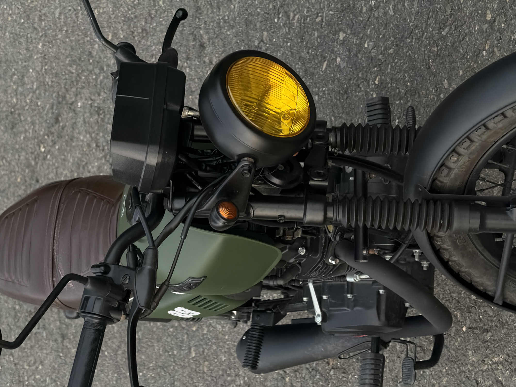 The image shows a close-up, high-angle view of a motorcycle. The motorcycle features a vintage design with modern elements, as evidenced by the yellow-tinted headlight, brown leather seat, and olive green fuel tank. The handlebars, controls, front fender, and engine components are predominantly black. The image does not contain any people or provide enough information to determine its specific location in Đà Nẵng, Vietnam, or any actions or interactions occurring. The environment is limited to the asphalt surface beneath the motorcycle.