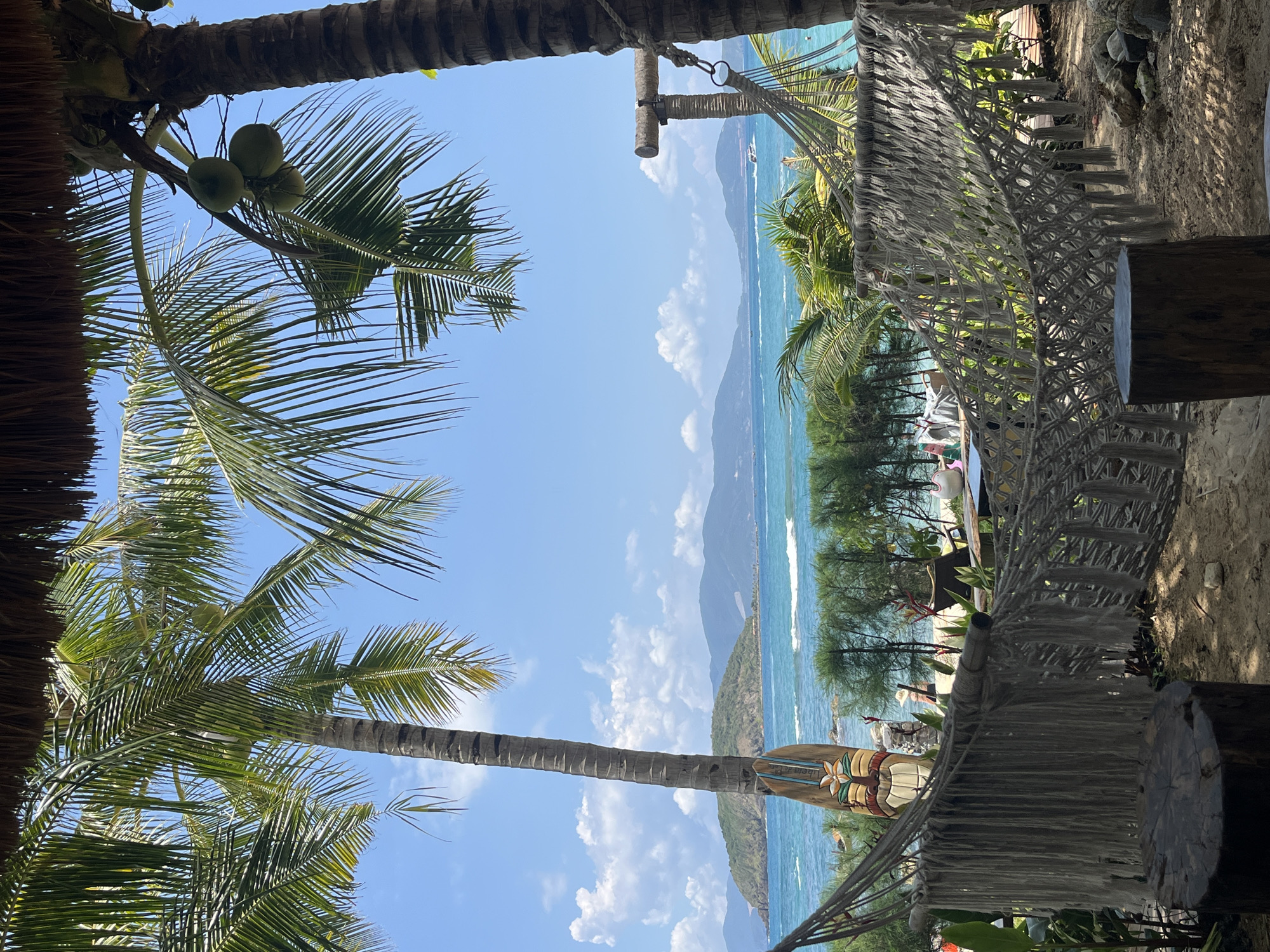 The image shows a coastal scene, likely a beach in Bac Nha Trang Ward, Vietnam. The view is framed by coconut palm trees and a thatched roof structure on the left, and a macrame hammock on the right. In the distance, there is a beach, some vegetation, and a blue body of water extending to a mountainous landmass under a sky with scattered clouds. The water appears to have gentle waves. No people are clearly visible.