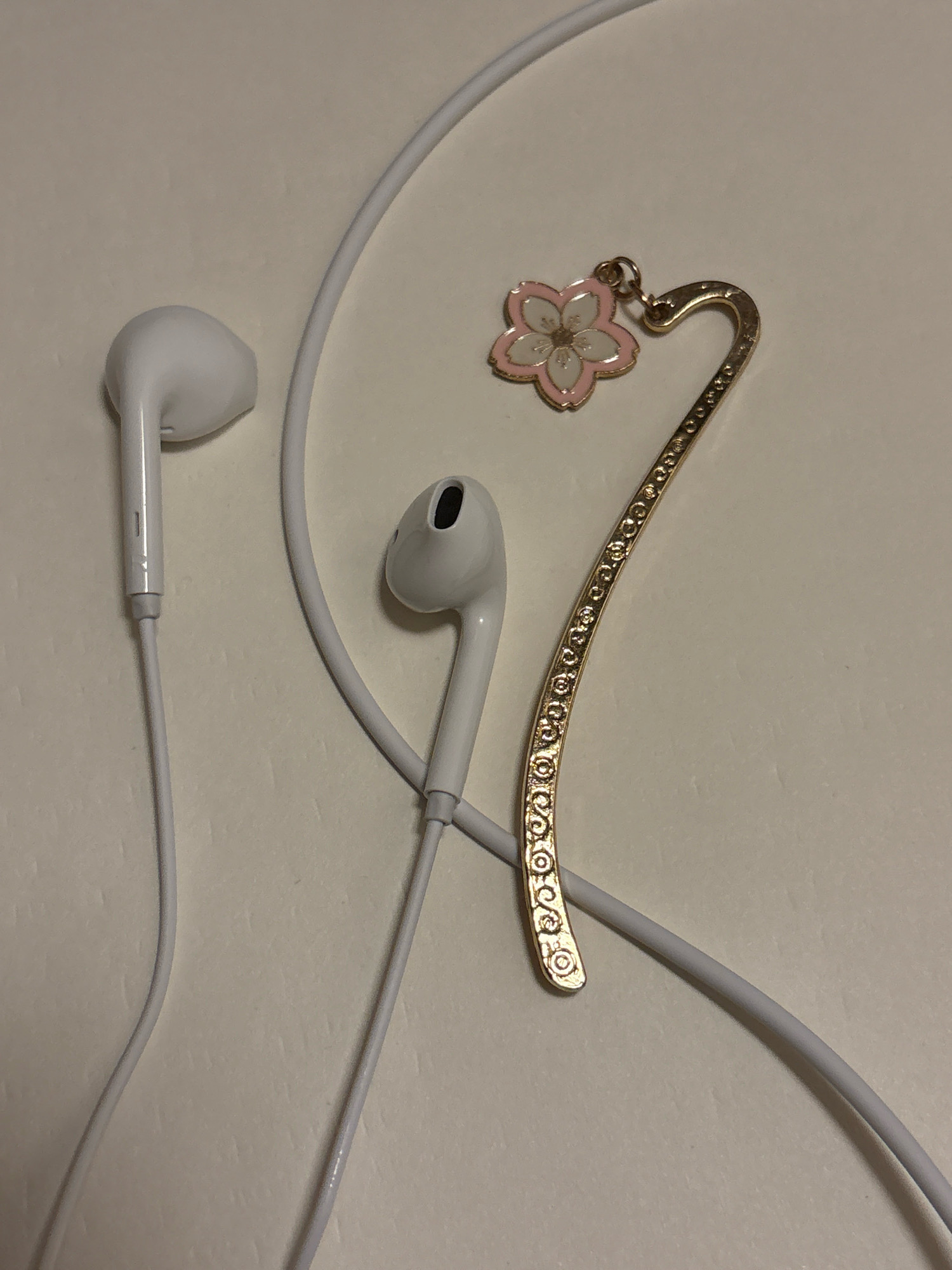 A pair of white wired earphones and a gold-toned metal bookmark with a pink and white flower charm lie on a light-colored, textured surface, possibly a desk or table. The earphones are positioned on the left side of the image, with the earpieces facing upwards. The bookmark is situated to the right of the earphones, curved and decorated with intricate spiral patterns. The flower charm, resembling a cherry blossom, is attached to the curved end of the bookmark. The background is plain and out of focus. The scene appears to be a still life arrangement of personal accessories.
