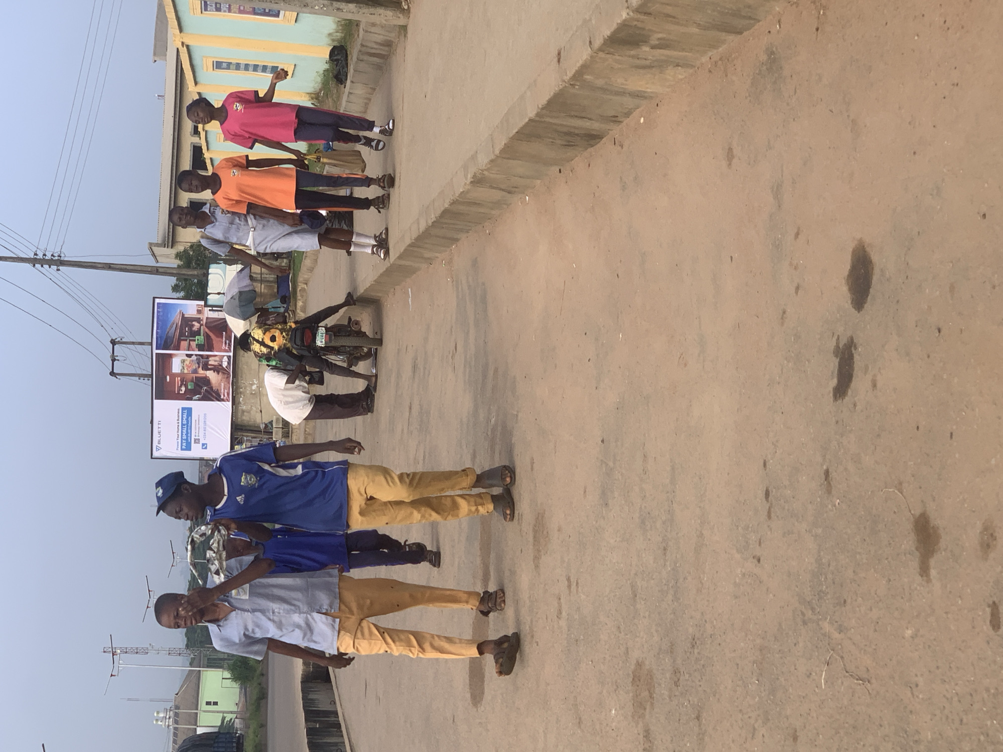 The image depicts a street scene in Shagamu, Nigeria, with several individuals walking on a paved surface. In the foreground, two young males are walking from left to right. The one on the left wears a light blue collared shirt and tan pants, holding a fan in his right hand near his face. The one on the right wears a blue and white sports jersey and tan pants, and appears to be holding something in his hands. Behind them, another person in a blue shirt and pants is visible. Further down the street, three young individuals are walking from left to right. The first is in a grey uniform with a white belt. The second wears an orange t-shirt and dark pants. The third wears a pink t-shirt and dark pants, carrying a brown paper bag. A motorcycle is parked on the left side of the image, with a person leaning over it. In the background, a large billboard displays advertisements, and structures identifiable as buildings and utility poles are present. The ground is paved and appears to have some dark stains. The sky is overcast.