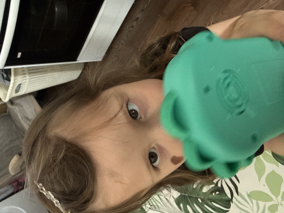 An indoor scene depicts a young child looking directly at the camera, partially obscured by a teal-colored object. The child has light brown hair, secured with a silver-colored barrette on the right side of her head (viewer's left), and a dark mole on her left cheek (viewer's right). She holds a rounded, possibly plastic or silicone toy with protrusions, obscuring her mouth and chin. The background on the left features a white surface with a prominent green tropical leaf pattern. In the mid-ground, a white, ribbed plastic laundry basket containing dark fabric is visible. To the right, a white kitchen oven with a dark glass door and a visible interior rack stands on dark brown wooden flooring, which also extends across the lower right portion of the frame. The specific location in the city of Кипчак, Russia, cannot be determined from the visual information presented.
