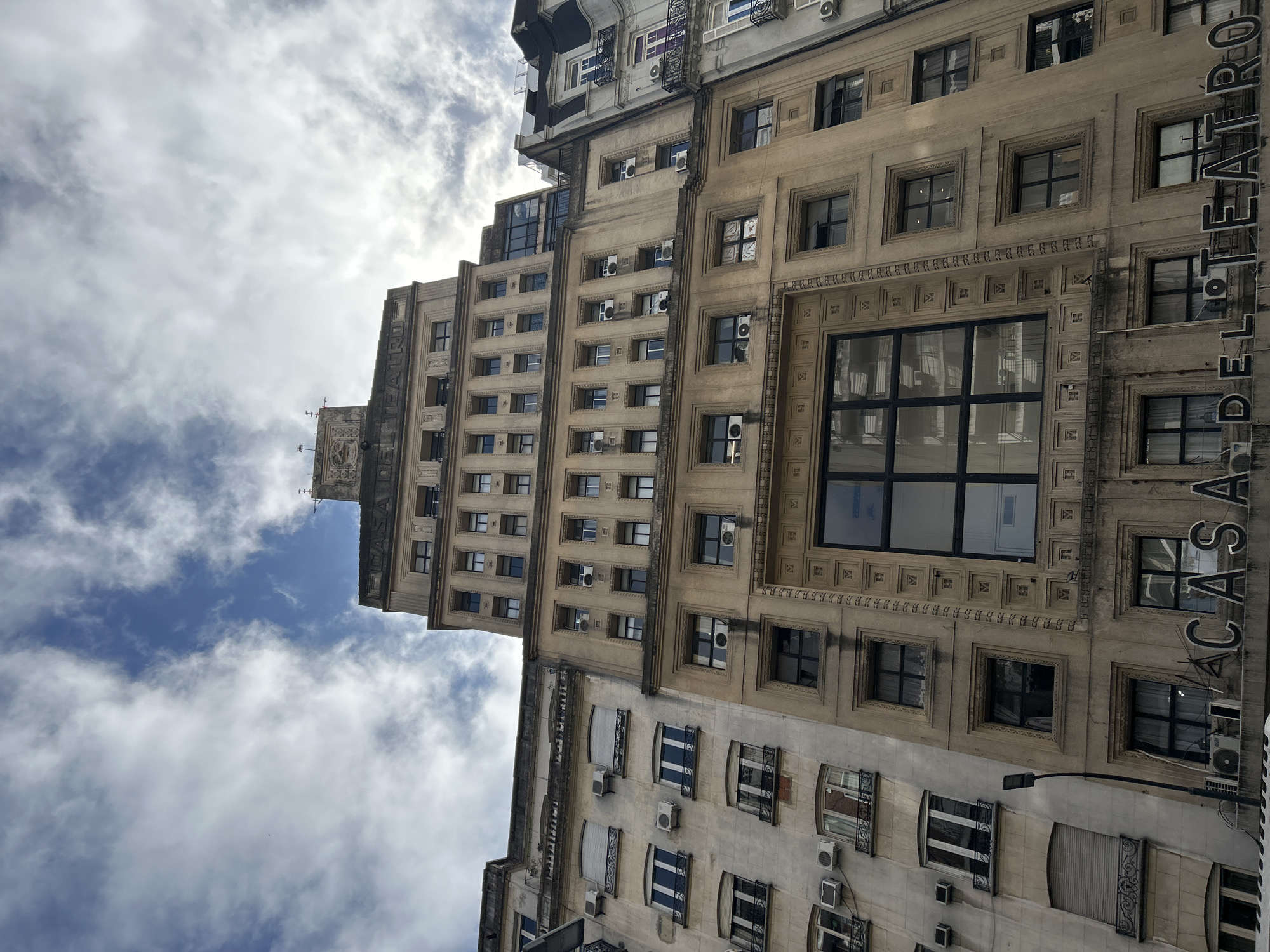 The photograph, captured from a low angle, looks up at the facade of the "Casa del Teatro" building in Buenos Aires, Argentina, under a cloudy sky with patches of blue. The architecture is characteristic of the early to mid-20th century, with a mix of Art Deco and classical influences. The building's facade is adorned with numerous windows, air conditioning units, and decorative moldings. The prominent signage, "CASA DEL TEATRO," is clearly visible at the lower part of the image. The surrounding buildings also display similar architectural styles, indicative of an urban setting. The overall impression is one of historical urban architecture, with a focus on the detailed design of the building.