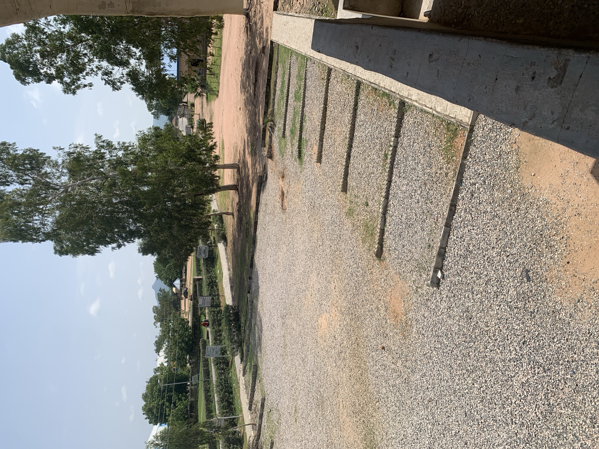 The image depicts an outdoor scene, likely on a university campus, in Jalingo, Nigeria. A large, gravel-covered area dominates the foreground, bordered by a concrete structure on the right and low, manicured hedges and trees in the midground. Several small, rectangular patches of grass, defined by concrete borders, are visible within the gravel area. Beyond the hedges, a dirt road leads into the distance, flanked by more trees and buildings with blue roofs. A few individuals, appearing as small figures, are seen walking on the road and near the hedges. In the background, a distant mountain range is visible against a hazy sky with scattered clouds. The overall impression is that of a sunny, dry environment.