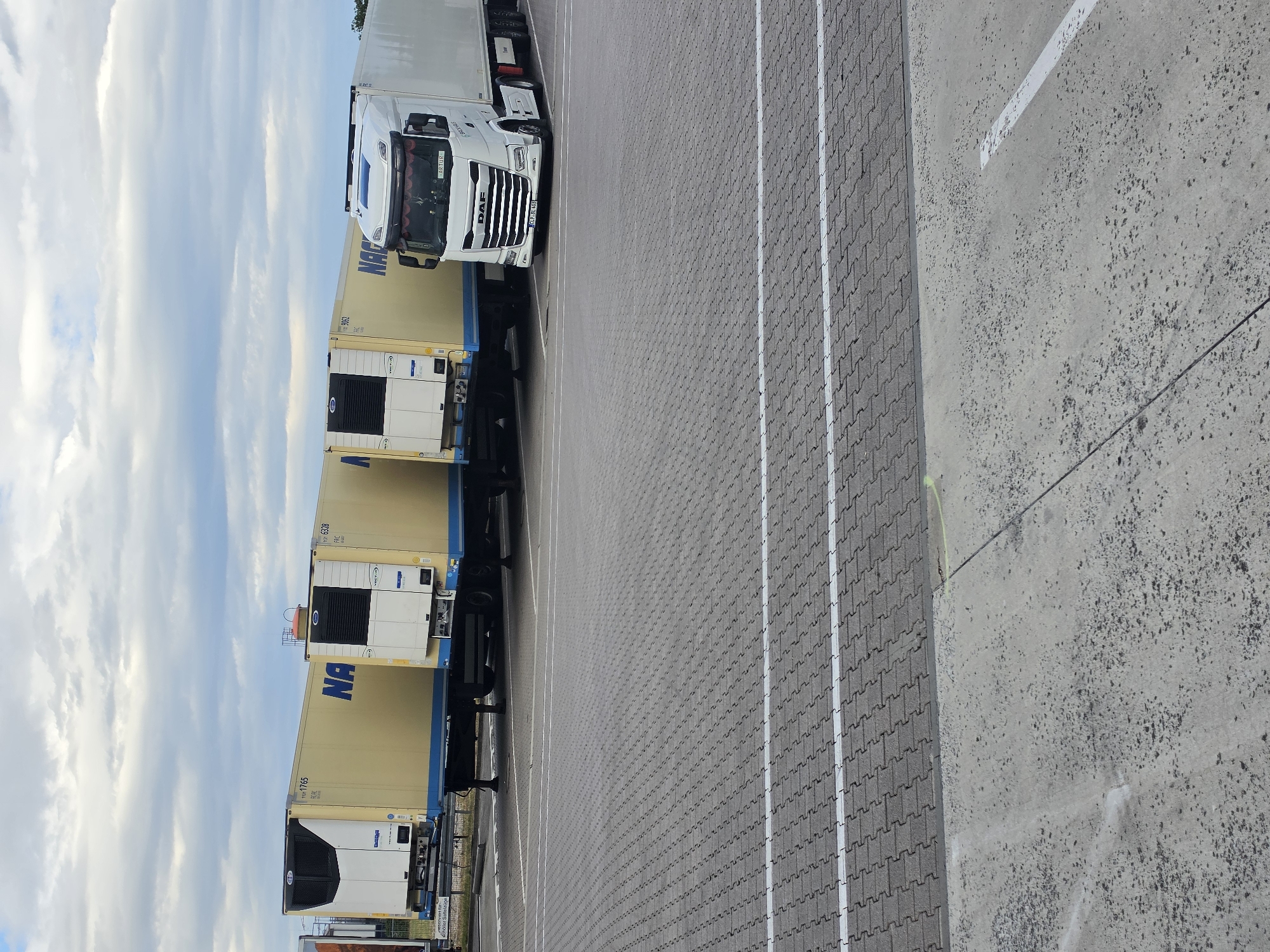 The image shows a parked DAF truck with two yellow shipping containers stacked on its trailer. The truck is white, and the containers are light yellow with blue lettering that reads "NA". One container has a white refrigeration unit on its front. The truck and containers are parked in a lot paved with gray interlocking bricks, marked with white lines. To the right of the paved lot is a concrete surface. The sky is visible in the background, filled with white and gray clouds against a blue sky, suggesting it is daytime. The scene appears to be a parking area, possibly at a logistics or distribution hub, given the presence of the commercial truck and shipping containers. There is no visible activity or people in the image. The license plate of the truck is visible, reading "JDR 2464". Text on the containers and a sign on the truck's side are difficult to read clearly. The overall impression is one of a static scene in an industrial or commercial outdoor setting. The image is taken from a slightly elevated, angled perspective, rotated 90 degrees clockwise.