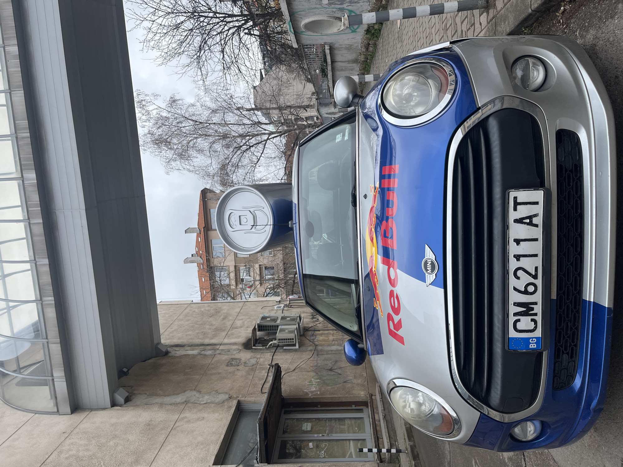 The image depicts a silver and blue Mini Cooper with a Red Bull logo and a large Red Bull can attached to the roof, parked on a street in Sofia, Bulgaria. The car's license plate reads "CM 6211 AT" with the Bulgarian flag. The background shows an urban setting with buildings, bare trees, air conditioning units on a wall, and a modern glass building. The scene appears overcast.
