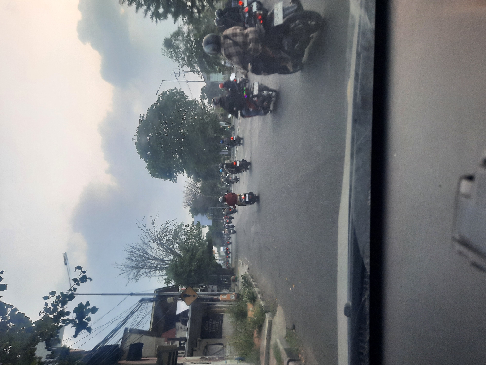 The image depicts a street scene in Buduran, Indonesia, likely during the late afternoon or early evening, given the soft lighting and overcast sky. The perspective is from inside a vehicle, looking forward through the windshield, which occupies the bottom portion of the frame. The road is paved and appears to be moderately trafficked with numerous motorcycles. Several individuals are riding motorcycles, some wearing helmets and casual clothing.
On the left side of the road, there are buildings, including a structure with signage and a yellow diamond-shaped warning sign on a pole. Vegetation, including trees and bushes, lines the side of the road and stretches into the distance. On the right side, more trees are visible. The sky is predominantly cloudy, with some lighter patches indicating diffused sunlight. The overall atmosphere is that of an urban or suburban street with active motor vehicle traffic.