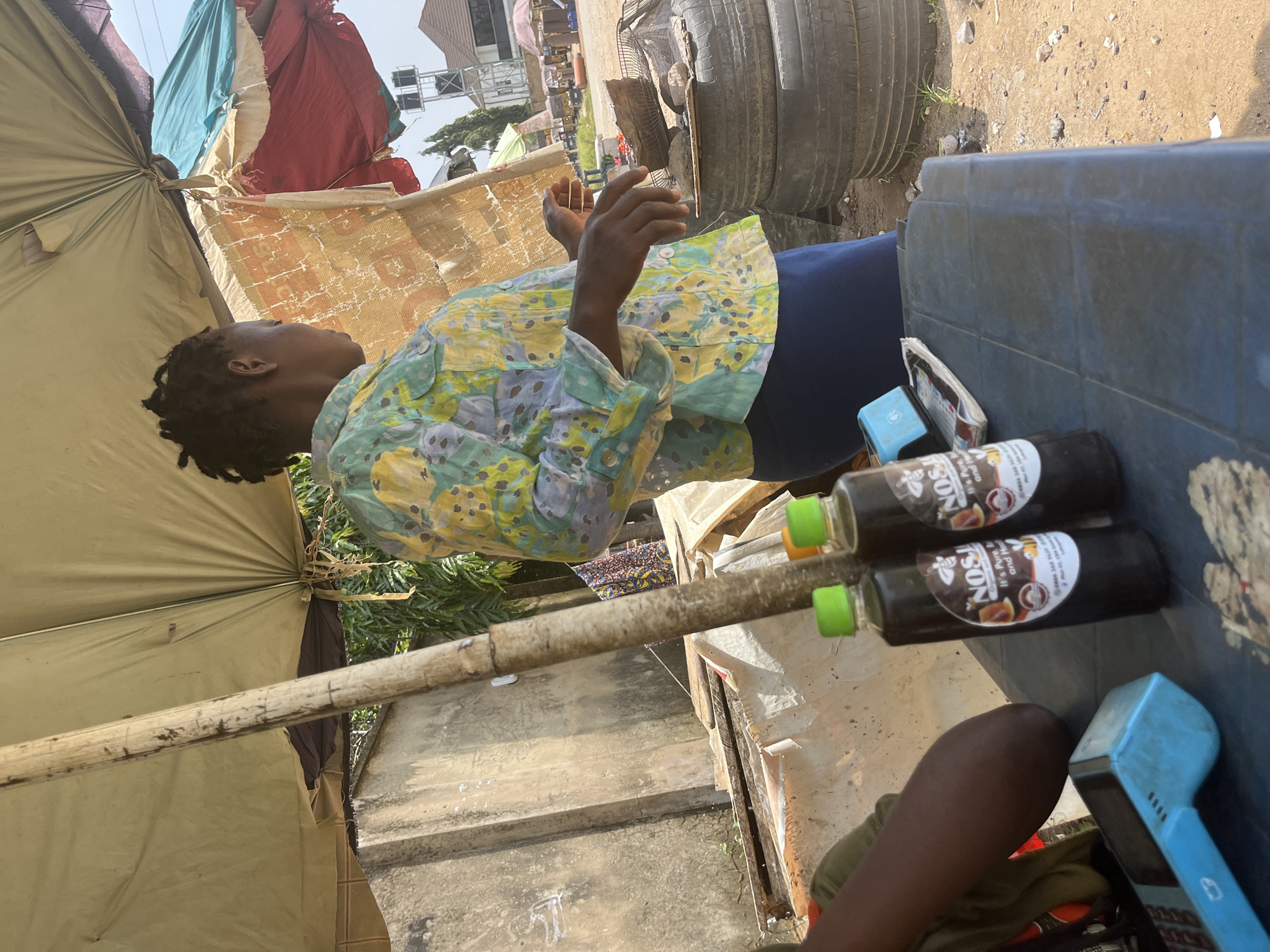 In Abuja, Nigeria, under a makeshift canopy, a Black woman wearing a colorful floral-patterned shirt and a dark blue skirt stands behind a blue plastic table. Two bottles of what appears to be a dark liquid, labeled "NOSA Organic Honey," are prominently displayed on the table next to a blue card reader. The woman is looking to her right, with her hands held in front of her. In the background, stacks of old tires are visible, along with a concrete wall and lush green foliage. Beyond that, a distant view of buildings and a water tower suggests an urban setting. The ground is covered in dirt and scattered stones.