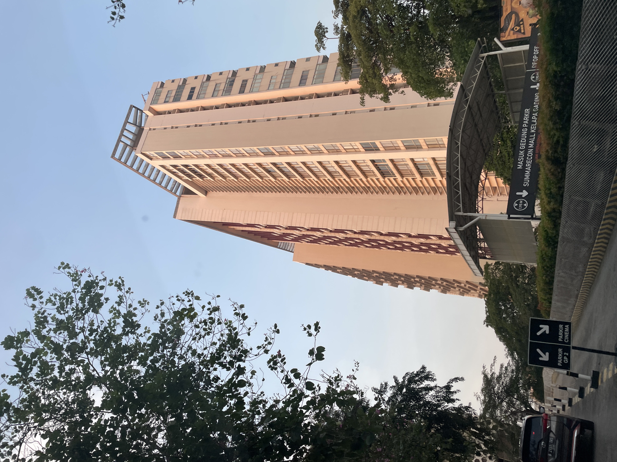 The image shows a tall, light-colored building, possibly a parking garage, with many windows and horizontal lines. The view is from a low angle, looking up at the building. A sign on the right indicates that the building is the "Summarecon Mall Kelapa Gading" parking garage, with a "Drop Off" area. A second sign indicates directions for "Parkir GP 2" and "Parkir Cinema". In the foreground, there are tree branches and a car. The picture appears to be taken outside the Summarecon Mall Kelapa Gading complex in North Jakarta, Indonesia. The license plate on the car shows "B 2131 TRS".