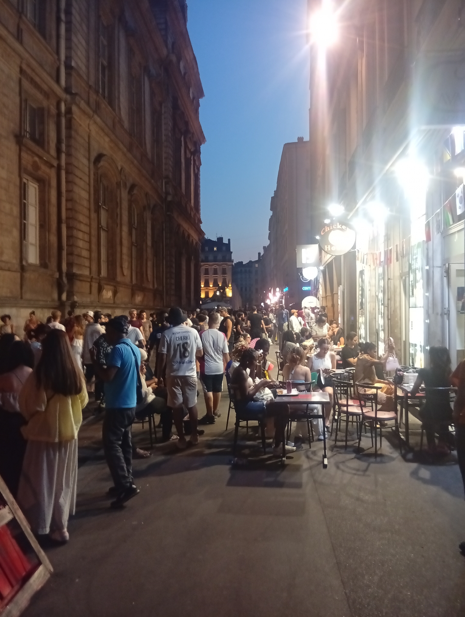 The image depicts a street scene at dusk in Lyon, France. A building with classical architecture, featuring multiple windows and ornate detailing, lines the left side of the narrow street. On the right, a brightly lit establishment with tables and chairs set up on the sidewalk suggests an outdoor dining area. Several people are visible, some standing and conversing in the street, while others are seated at the tables, engaged in dining or socializing. The sky is a deep blue, indicating late evening or early morning. The presence of lights and the atmosphere suggest a lively urban environment.