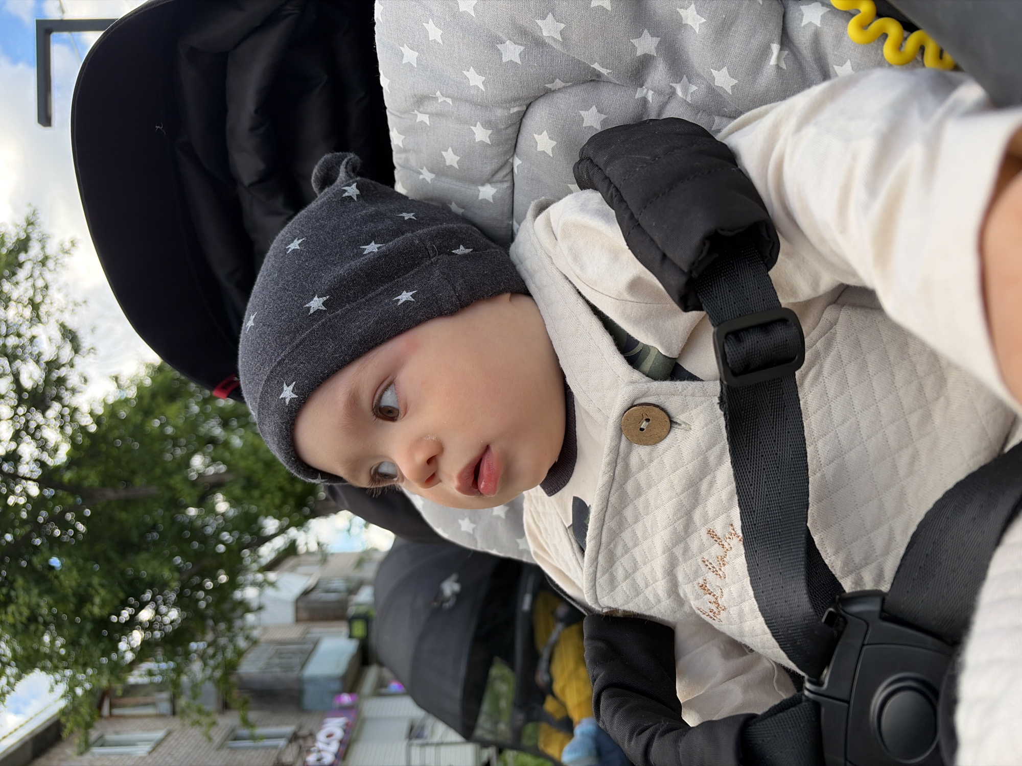 A baby is seated in a stroller in what appears to be an urban environment. The baby is wearing a dark gray beanie with white stars, a cream-colored quilted jacket with a wooden button, and is secured in the stroller with black safety straps. The stroller features a gray cushion with white stars and a black sunshade.
In the background, a tree with green foliage is visible, as well as a multi-story building with windows and a sign with Cyrillic letters. Based on the Cyrillic script, the location is potentially Chaikovsky, Russia.