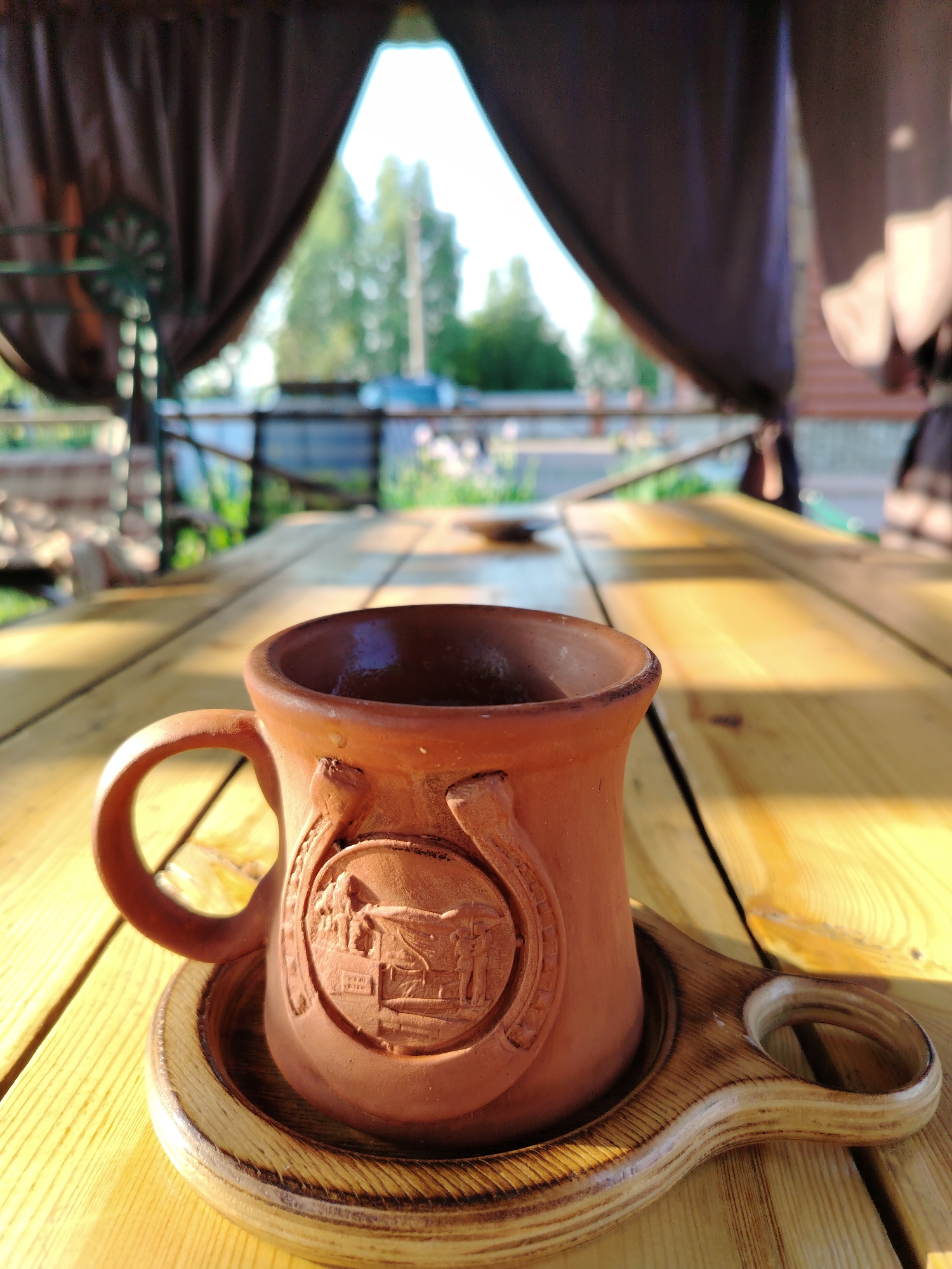 A close-up, low-angle shot captures a rustic terracotta mug with a horseshoe-shaped relief on a wooden table. The mug rests on a wooden saucer with a handle. Sunlight casts strong shadows and highlights across the wooden surface. In the background, out of focus, is a gazebo with draped fabric and a glimpse of greenery and trees, suggesting an outdoor setting, possibly in the city of Рассвет, Russia. The overall impression is one of a tranquil, sunlit moment.