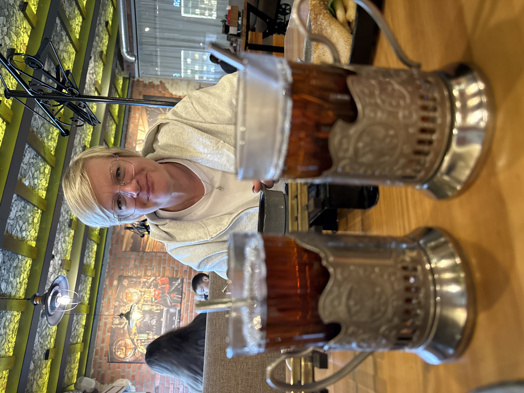 A woman with short blonde hair and glasses is seated at a table, looking towards the camera and smiling. She is wearing a light gray hooded jacket over a white t-shirt, with a silver necklace. In the foreground, two glasses of dark liquid, likely tea, are presented in decorative metal holders. The glasses are filled to the brim, with spoons in each. The table appears to be made of wood. In the background, a brick wall with framed artwork is visible, along with a ceiling with exposed beams and decorative lighting. Other patrons are seated at tables in the distance, with one individual wearing dark hair visible in the mid-ground to the left. The overall setting suggests an indoor dining or cafe environment.