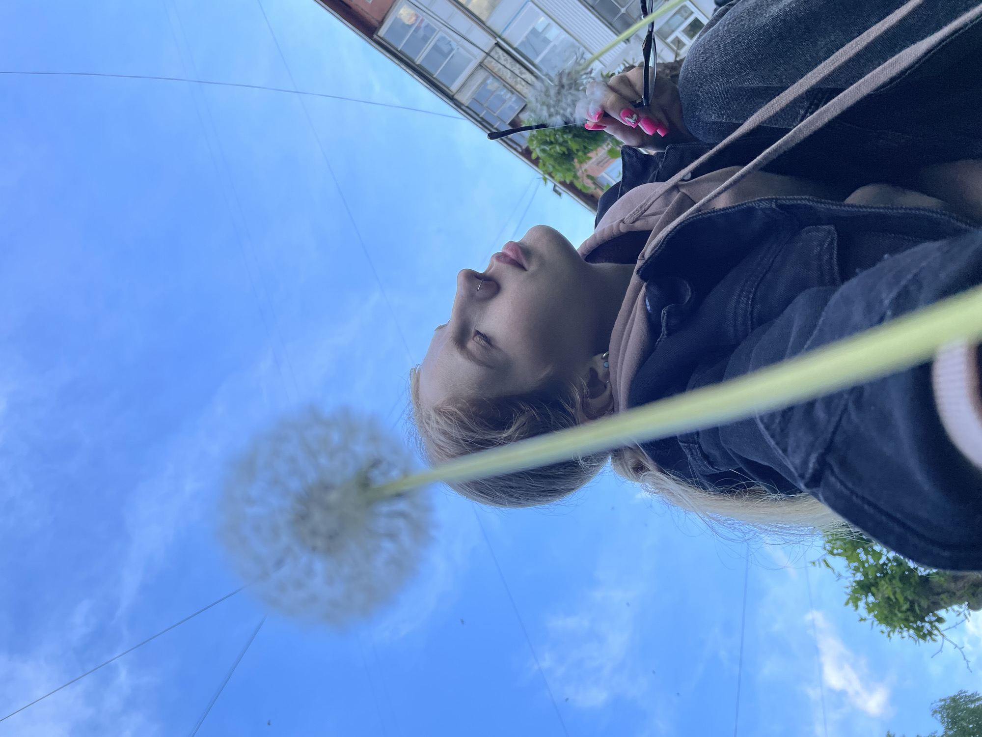 A low-angle shot reveals a person holding a dandelion in Izhevsk, Russia. The person is seen from the chest up, wearing a dark denim jacket over a light-colored hoodie. In their right hand, they are holding eyeglasses with bright pink fingernails. The focus is on the person's face and the dandelion head, with the sky as the backdrop. The dandelion stem aligns vertically with the person's face. The blue sky has sparse clouds. In the upper portion of the frame, a building with windows and some greenery can be seen. Small black dots, likely insects, are scattered across the sky.