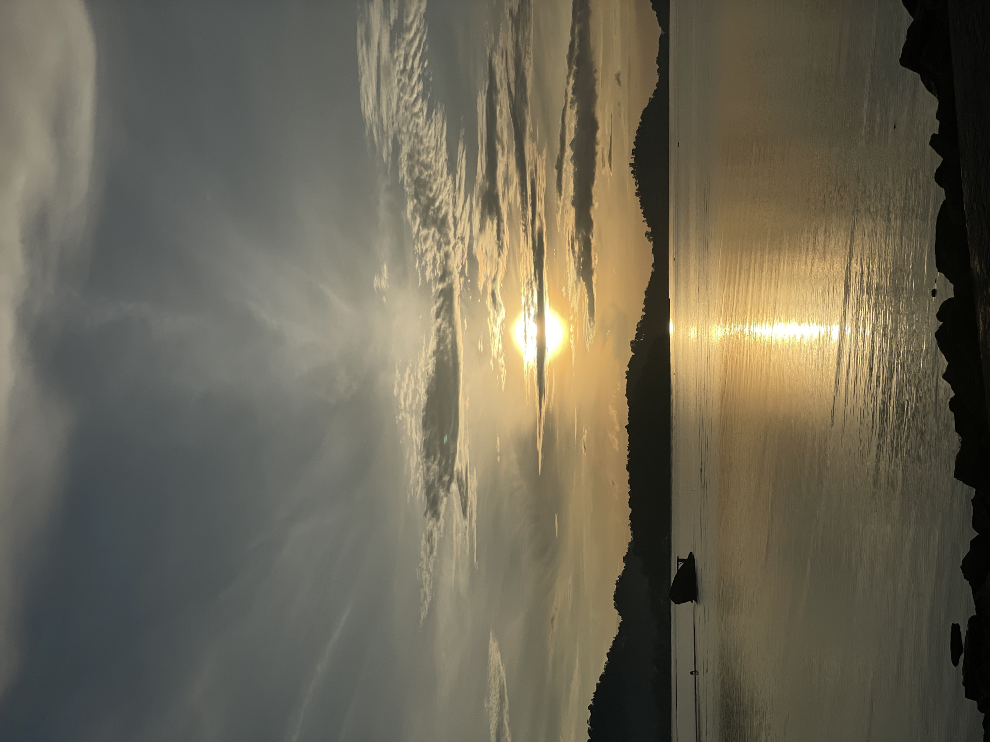 The image depicts a serene sunset over a body of water, likely in Phuket City Municipality, Thailand. The sun, a bright orb, is partially obscured by scattered clouds, casting a golden glow across the sky and reflecting on the water's surface. Silhouetted against the horizon are the lush, rolling hills of the coastline. In the foreground, dark rocks line the edge of the water. A solitary, dark-colored boat is visible on the water, moving from left to right. The overall atmosphere is tranquil and picturesque, characteristic of a coastal sunset.