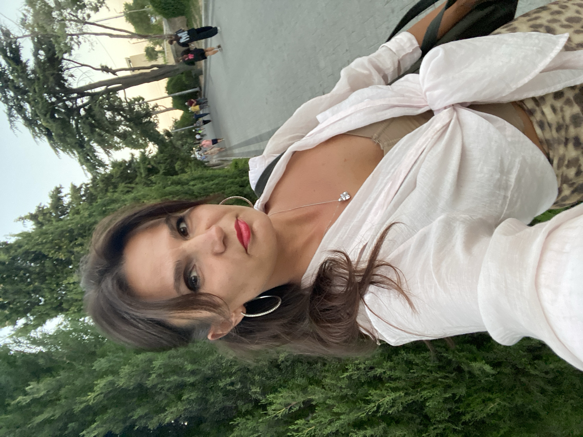 In the city of Feodosia, Ukraine, a woman stands on a paved path lined with cypress trees. She is taking a selfie, facing the camera with a serious expression. She has dark, wavy hair pulled back, large hoop earrings, and is wearing a light pink, loosely tied shirt over a beige camisole. Her lower body is adorned with a leopard-print skirt. In the background, several people are walking along the path, and tall trees form a verdant backdrop against a bright sky.