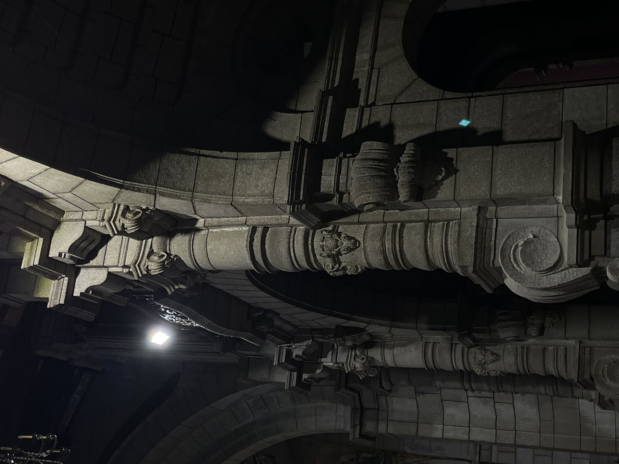 The image displays an interior view of an old stone building, possibly a church or historical structure, in Mérida, Venezuela. The architecture features prominent stone arches, massive columns with intricate carvings, and stone block walls. A bright, square light source is visible in the upper left quadrant, illuminating a section of the ornate architecture. Shadows are cast heavily across the scene, suggesting dim lighting conditions, characteristic of an evening or dimly lit interior. There are no people visible in the frame. The overall impression is one of grandeur and historical significance, with the stone elements and architectural style pointing towards a European influence common in colonial-era buildings in Latin America.