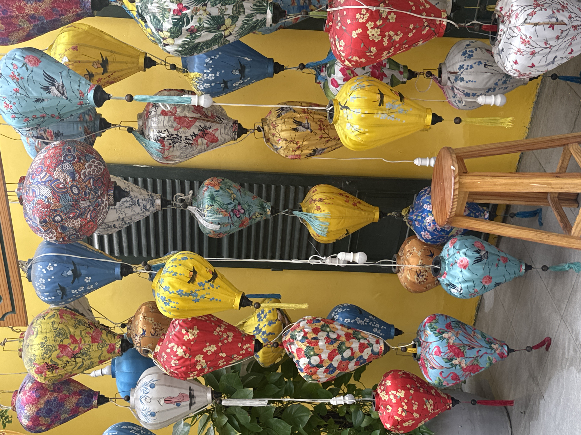 This image depicts a vibrant display of numerous Vietnamese silk lanterns hanging in front of a yellow wall, characteristic of a street vendor or shop in Nha Trang, Vietnam. The lanterns are of various sizes and colors, adorned with intricate traditional patterns, including floral motifs, birds, and scenes. Many lanterns feature tassels and decorative beads.
The scene includes several hanging lights, some connected to the lanterns. A small wooden stool with a round seat is placed in the foreground, suggesting it is part of the display or for customers. To the left, a large leafy plant is visible, adding a touch of greenery. The ground is paved with grey tiles. The overall impression is one of a bustling marketplace or a shop specializing in decorative Asian lanterns. No people are present in the image.