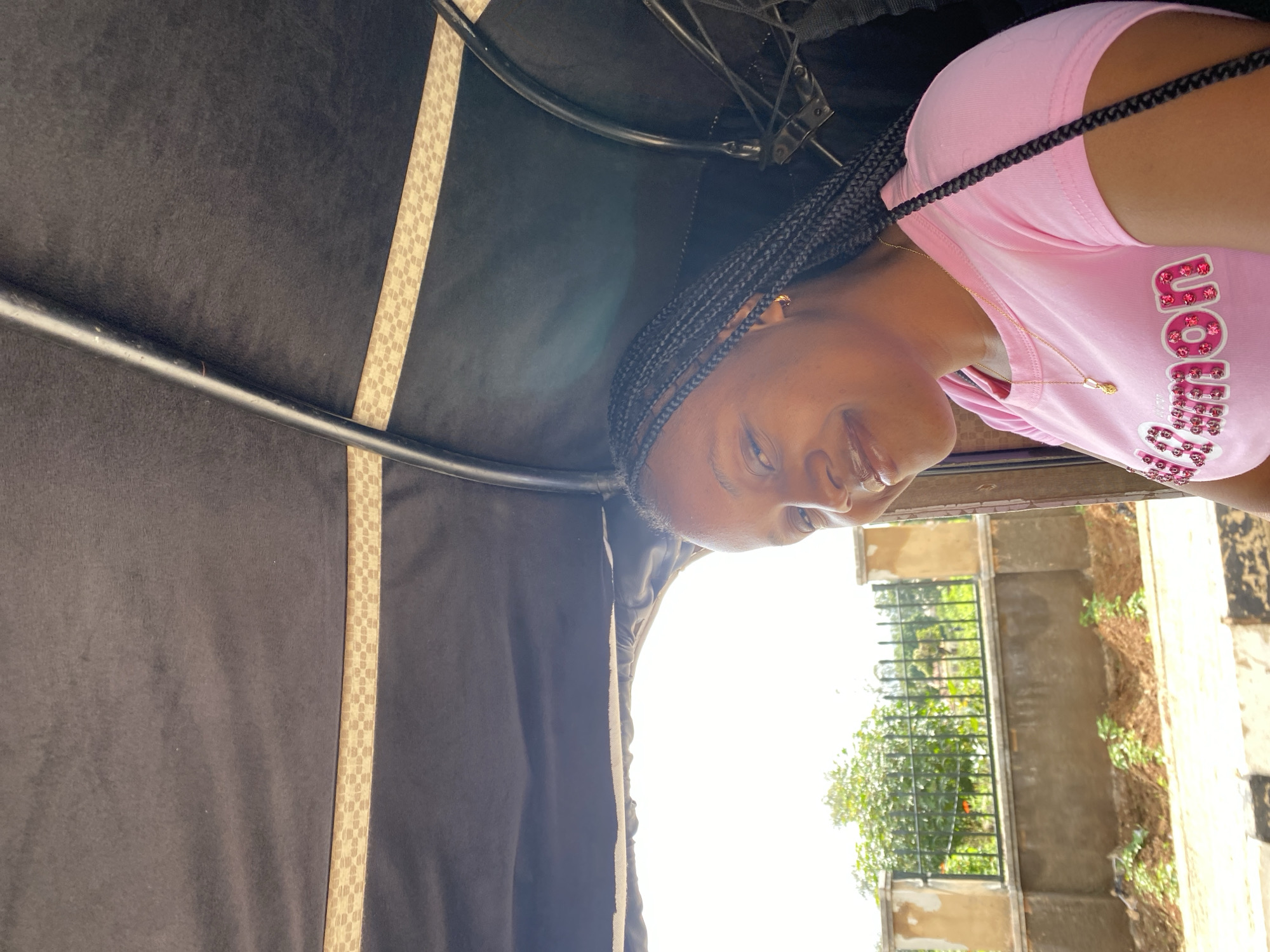A young Black woman with braided hair is inside the covered passenger area of a motorized rickshaw, likely a Keke Napep, in Bar Jirgi Summa, Nigeria. She is wearing a bright pink t-shirt adorned with the word "Pokemon" in large, sparkling pink letters. A delicate gold chain with a small pendant rests on her collarbone. Her head is turned slightly to her right, and she is smiling. Through the open side of the rickshaw, a fence is visible, beyond which are trees and buildings. The light suggests it is daytime. The interior of the rickshaw is dark, covered by a black fabric canopy, with some structural elements and a patterned trim visible.