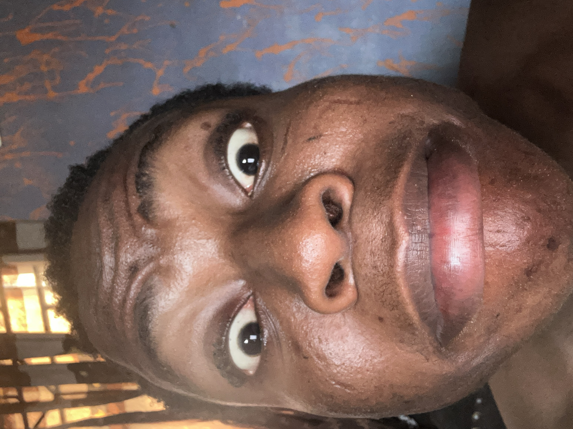 A close-up, tilted view of a person's face with brown skin. The subject has dark eyes, a prominent nose, and lips that are parted. The background is a mix of dark and light colors, with some orange streaks. It is not possible to determine the location or any actions or interactions from the image.