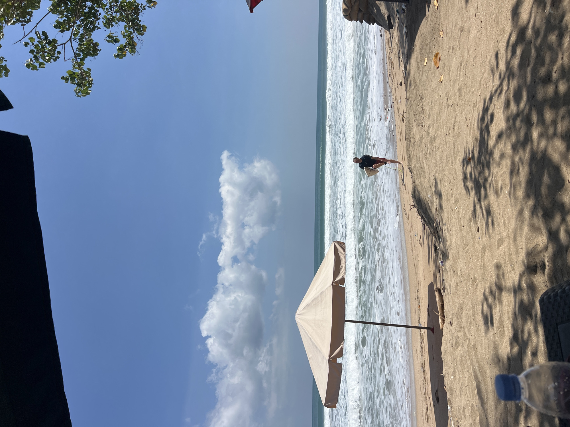 A lone surfer walks along the shore of Kuta Beach, Indonesia, carrying his surfboard. The sky is a vibrant blue with scattered white clouds. Waves crash onto the sandy beach, and a beach umbrella offers shade. In the foreground, a plastic water bottle sits on a table, partially obscuring the view of the sand and shadows cast by nearby trees. The overall scene depicts a sunny day at a popular tourist destination.