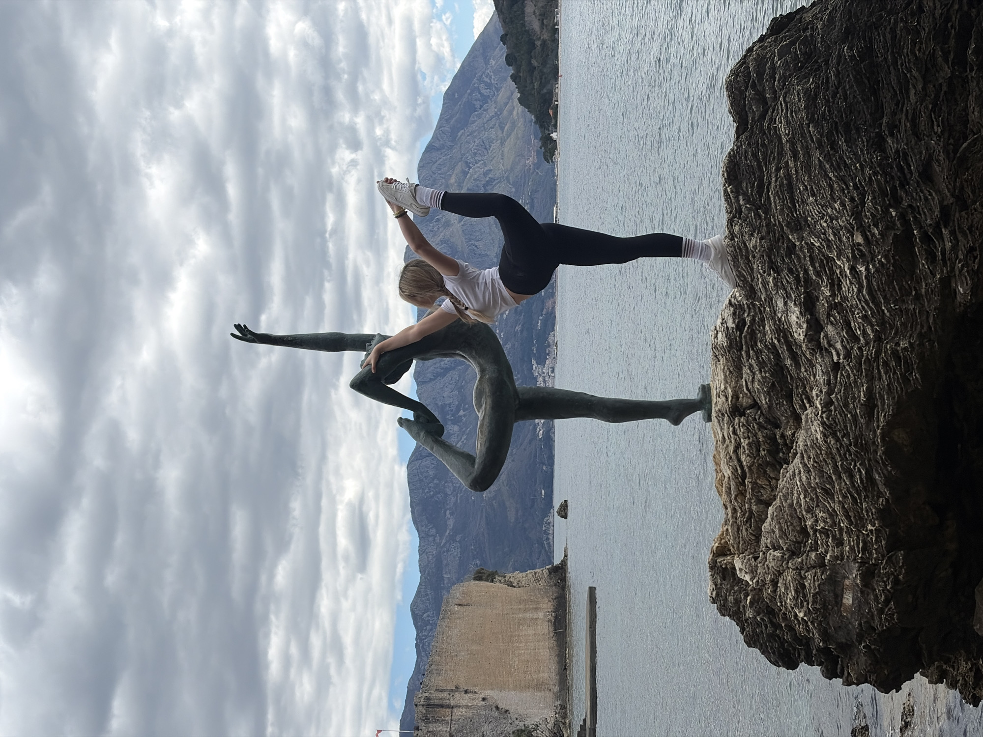 A rotated wide shot shows a young woman in a yoga pose atop a seaside rock formation, interacting with a metal sculpture, with a mountainous background and ocean in the city of Budva, Montenegro. The woman is fair-skinned with blonde hair in a braid, wearing a white t-shirt, black leggings, white socks with stripes, and white sneakers. She is holding her right foot and her left leg is extended straight out. She has one hand touching the sculpture. The sculpture features two human-like figures intertwined in a dancing pose. The rock formation is dark and textured. The sea is calm and stretches to the horizon. Mountains are in the distance under a cloudy sky. The shot is at an angle, rotated ninety degrees clockwise.